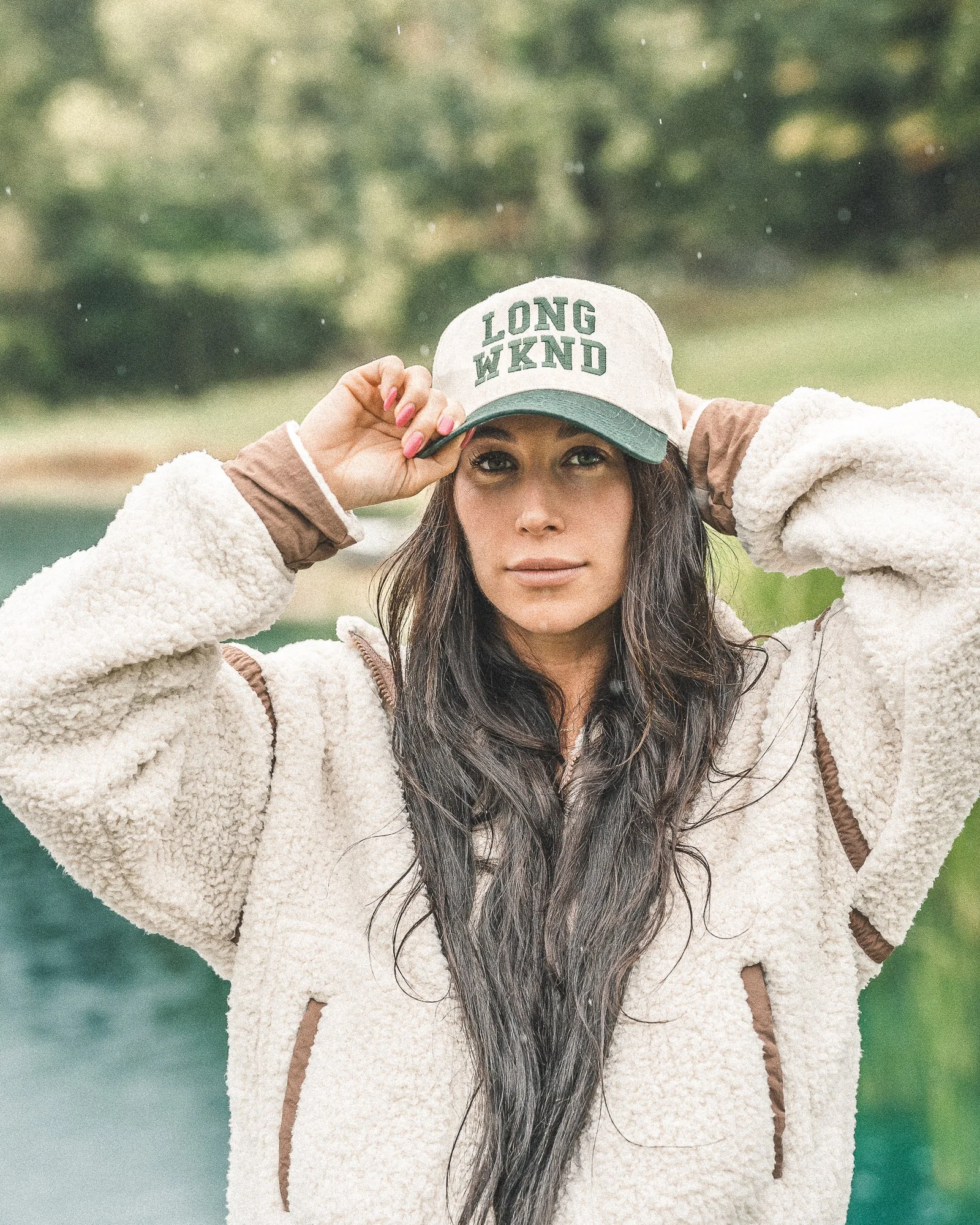 A woman with long dark hair wearing a beige fleece jacket and a gray cap that says "LONG WKND" poses outdoors in front of a body of water and green trees, holding the brim of her cap with both hands.