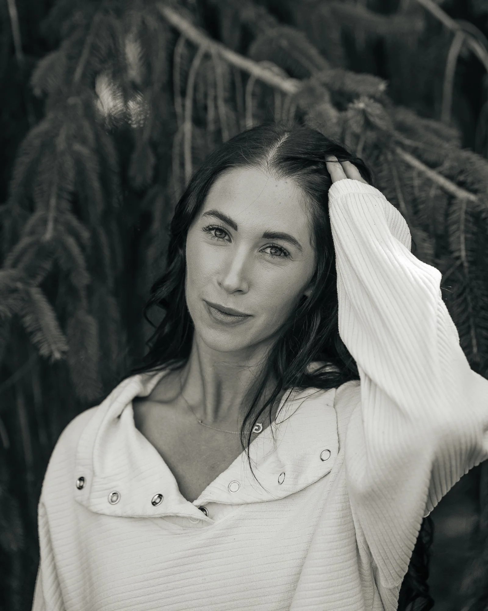 A woman with dark hair and light skin poses outdoors, touching her hair with one hand, standing in front of pine trees.