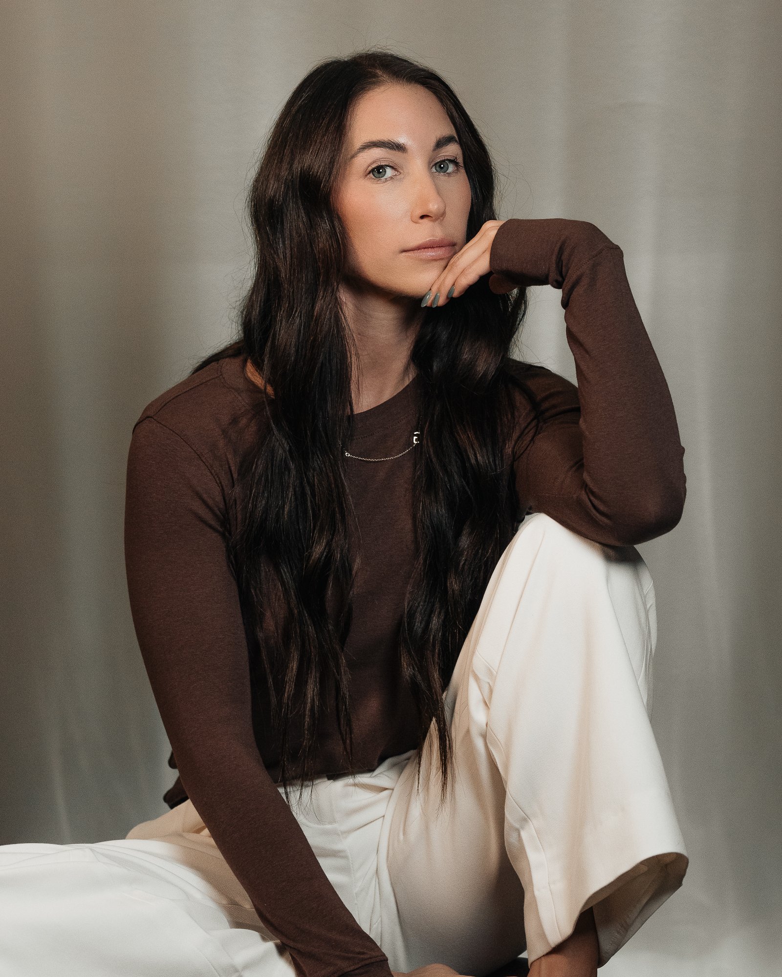 A woman with long dark hair, wearing a brown long-sleeved shirt and white pants, sitting with her chin resting on her hand against a plain background.