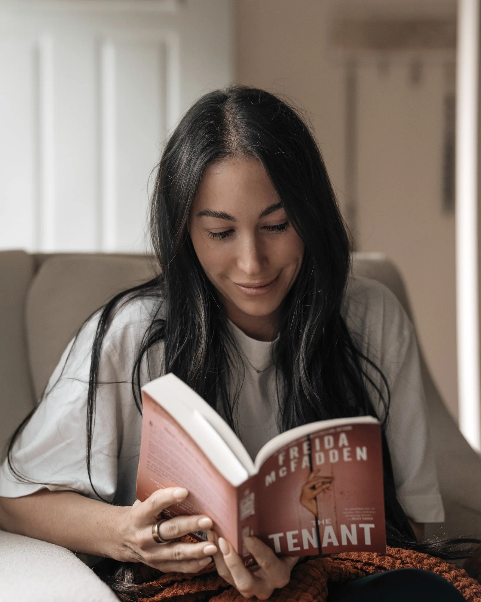 Woman with long dark hair reading the book "The Tenant" by Freda McFadden while sitting on an armchair.