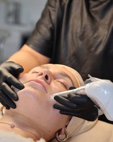 A woman receiving a facial treatment in a spa or clinic setting, with a professional wearing black gloves applying skincare products or facial tools to her face.