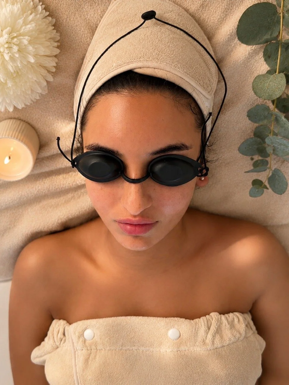 Woman lying down with spa goggles on, beige towel wrapped around her chest, beige headband, surrounded by a candle, a white flower, and eucalyptus leaves.