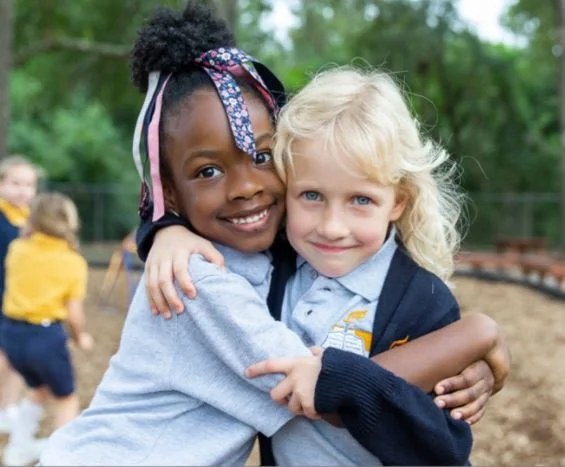 Two young girls hugging outdoors at school playground, smiling at the camera, with other children in the background.