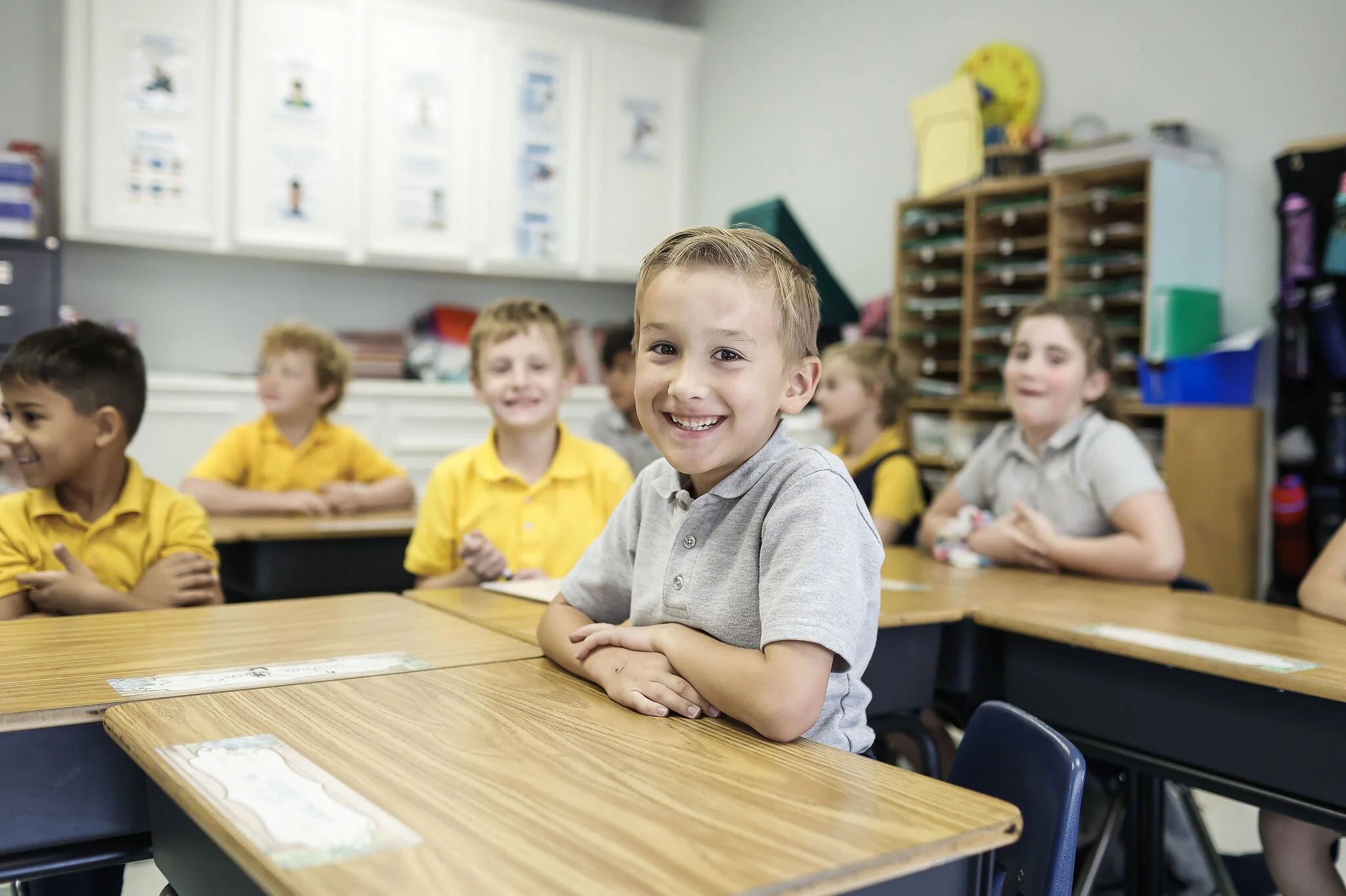 A group of young children sitting at desks in a classroom, smiling and looking at the camera, with a boy in a grey shirt in the foreground.