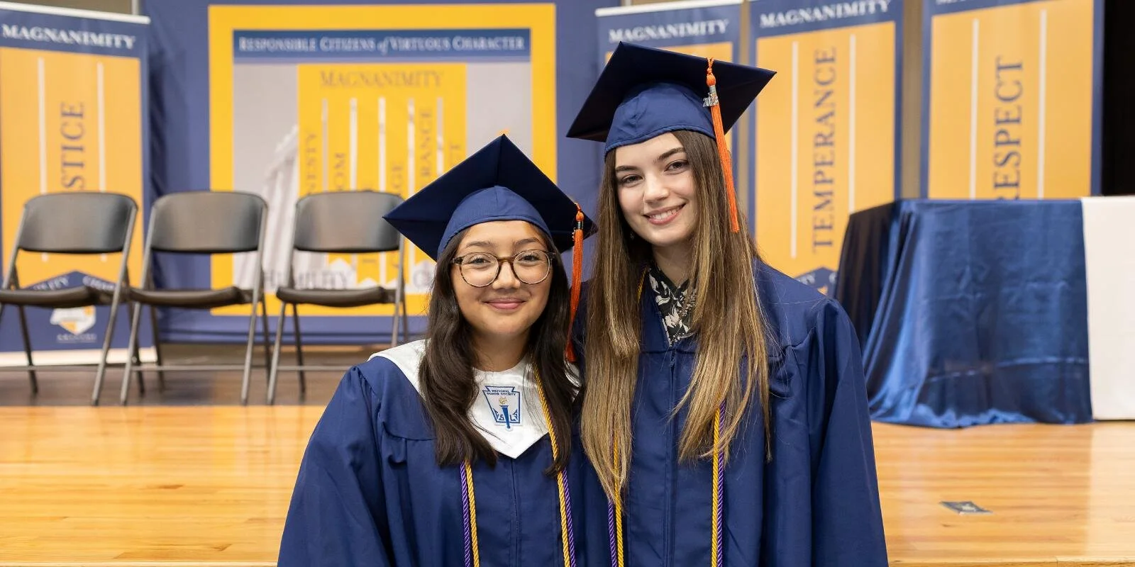Two young women in blue graduation caps and gowns smiling, standing on a wooden stage with chairs behind them and educational posters on the wall.