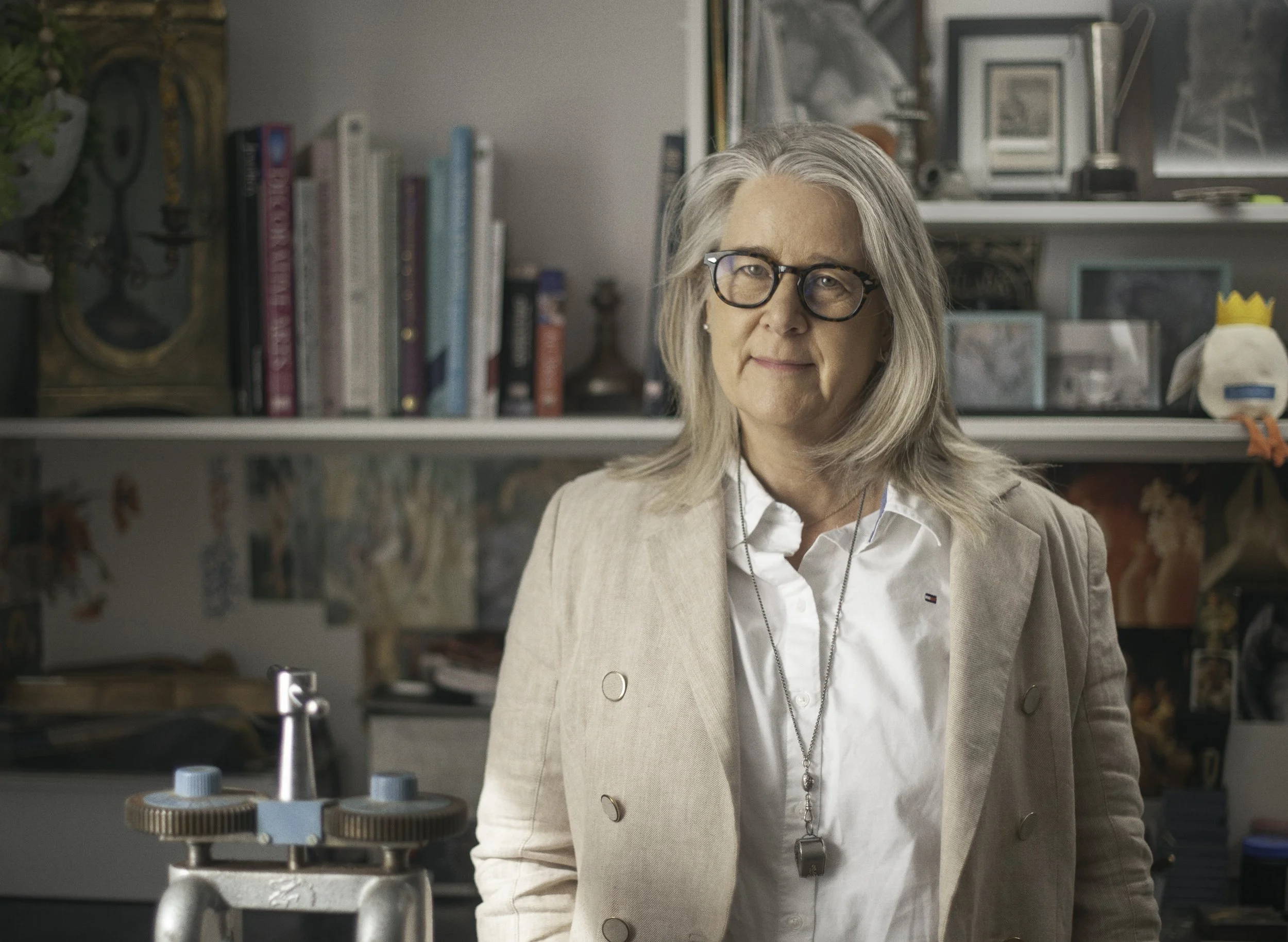 A woman with gray hair, glasses, and a white blouse with a beige blazer stands in an art or science room, with shelves of books, framed pictures, and art supplies in the background.