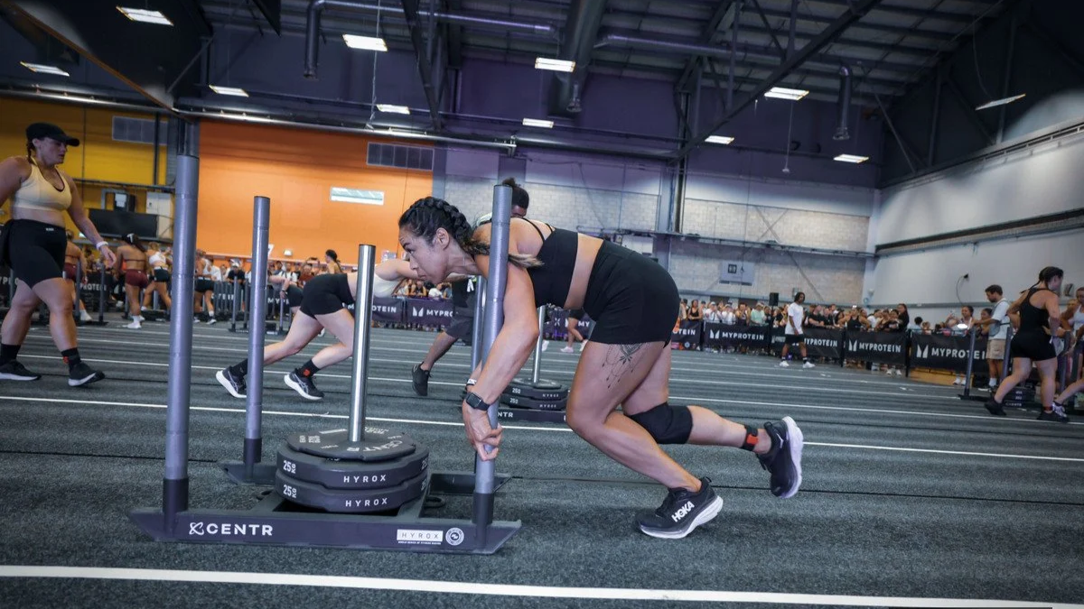 A woman participating in a fitness competition, pushing a weighted sled forward on a gym floor with other competitors and spectators in the background.