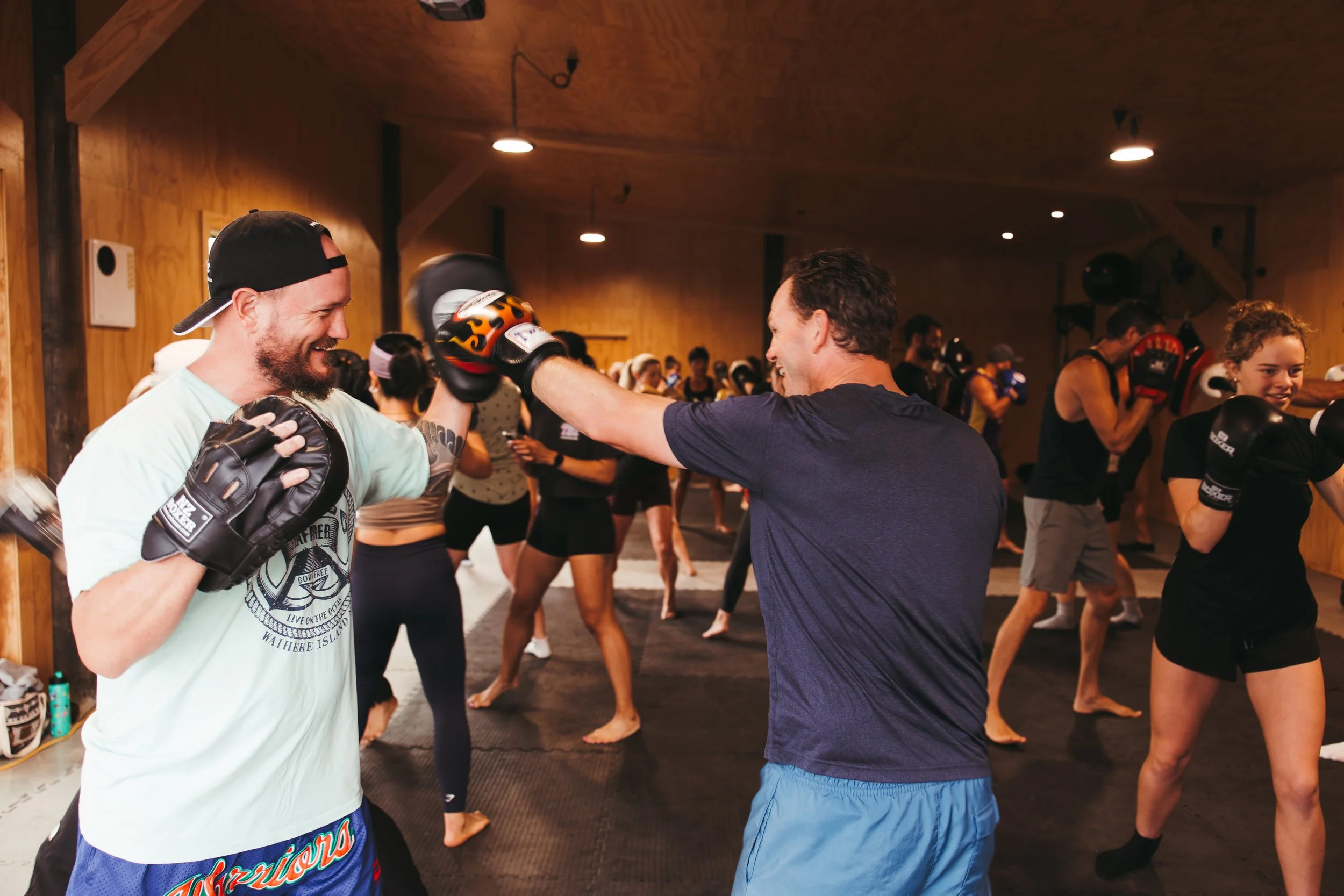 Two men practicing boxing at a boxing gym, doing drills with focus mitts, surrounded by other people dressed in athletic clothing in the background.