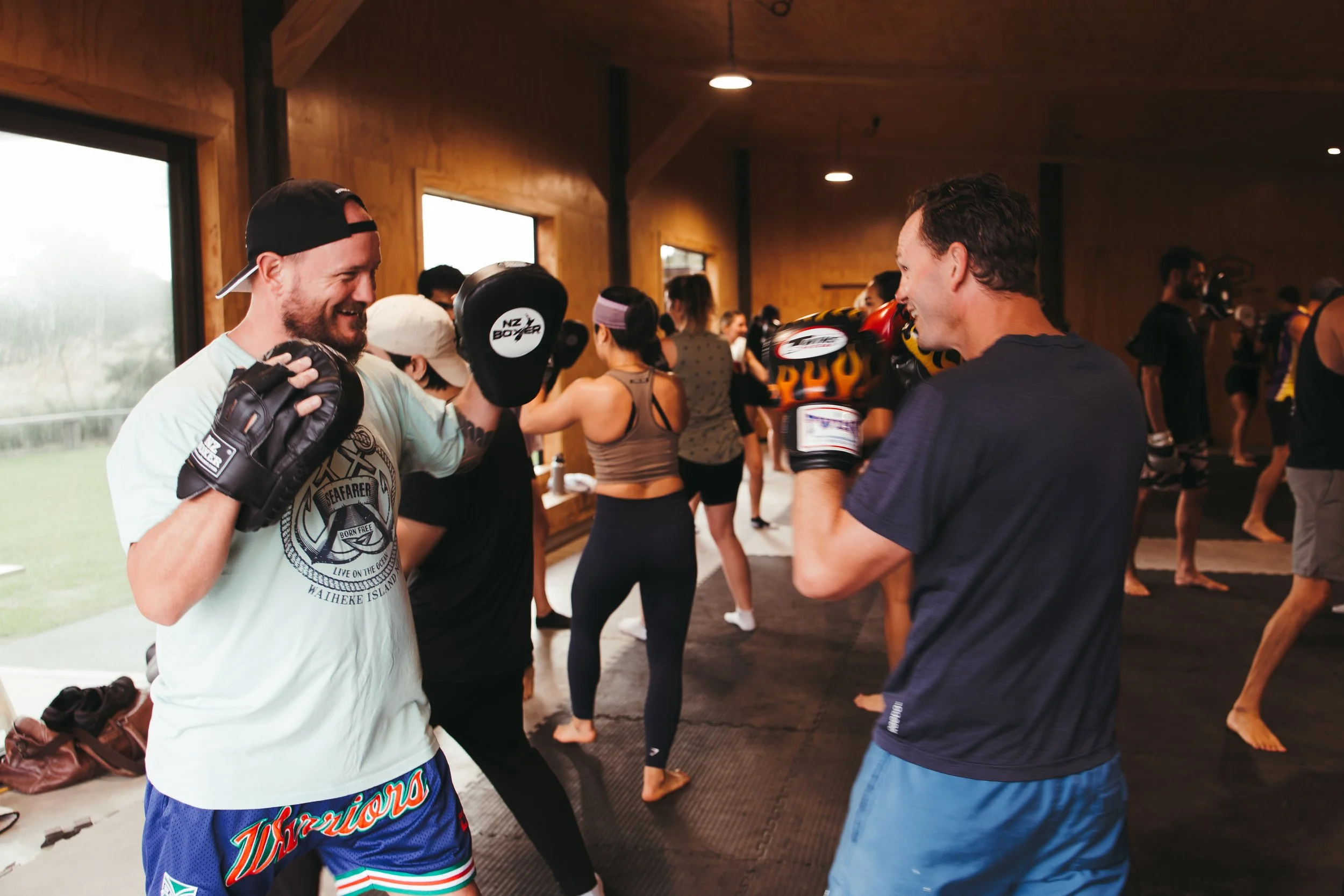 Two men practicing boxing in a martial arts gym with onlookers in the background.