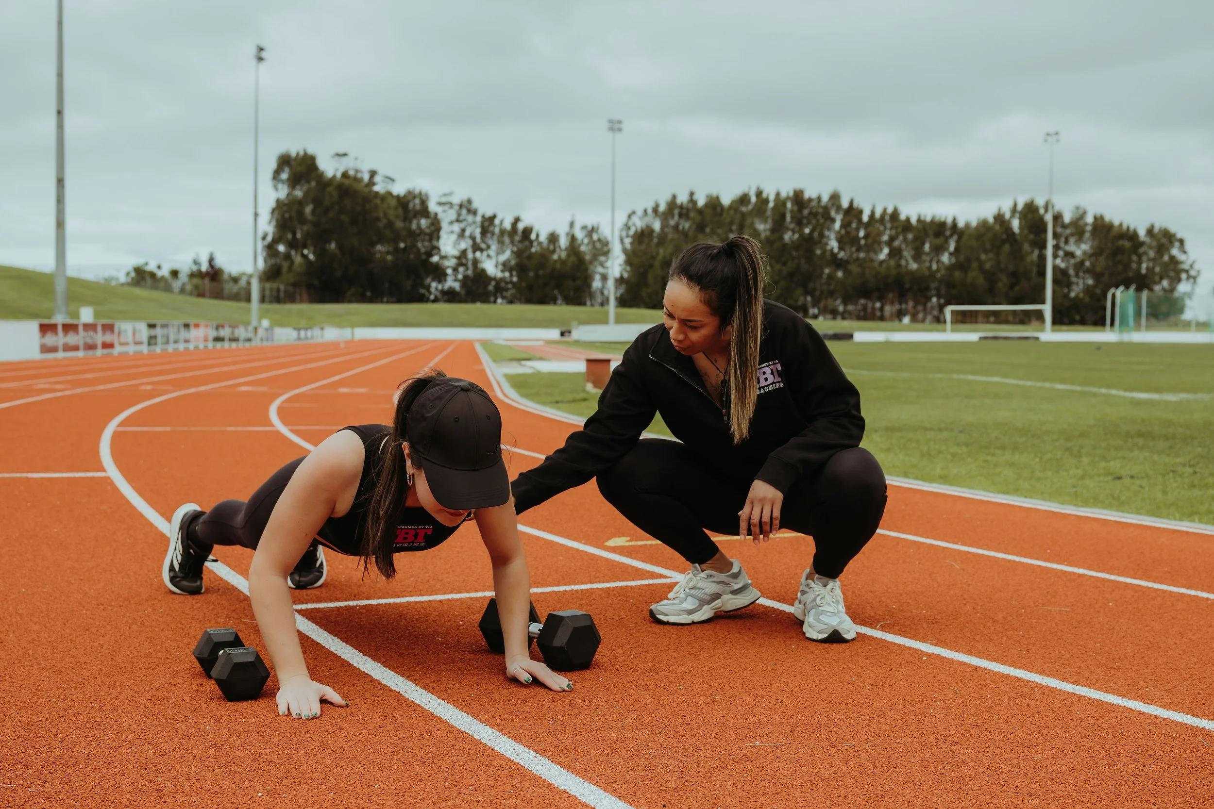 A woman coaching a young girl during track practice on a running track, with a cloudy sky and trees in the background.