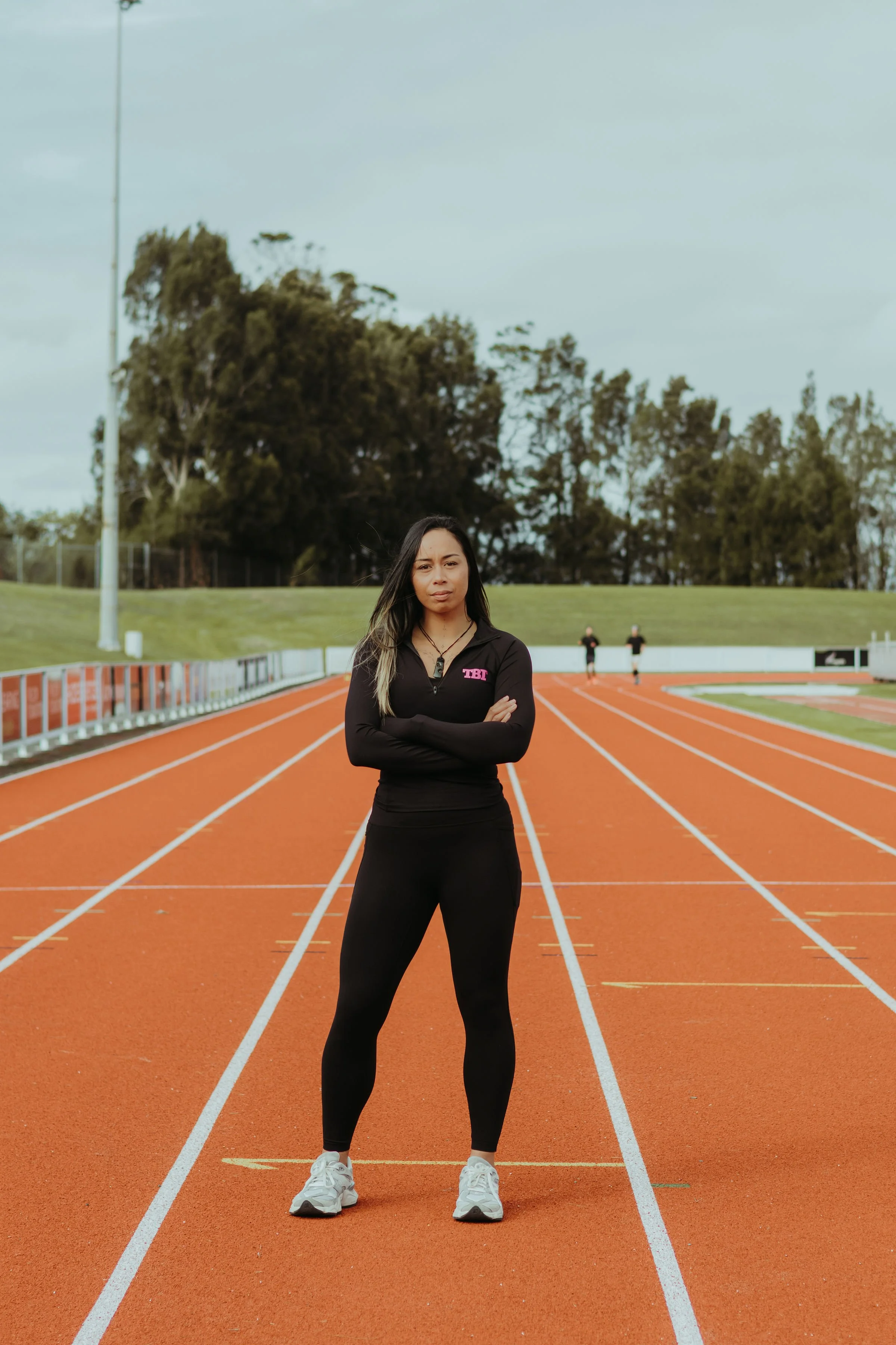 A young woman standing confidently with arms crossed on a running track at an outdoor athletic field, with trees and a cloudy sky in the background.