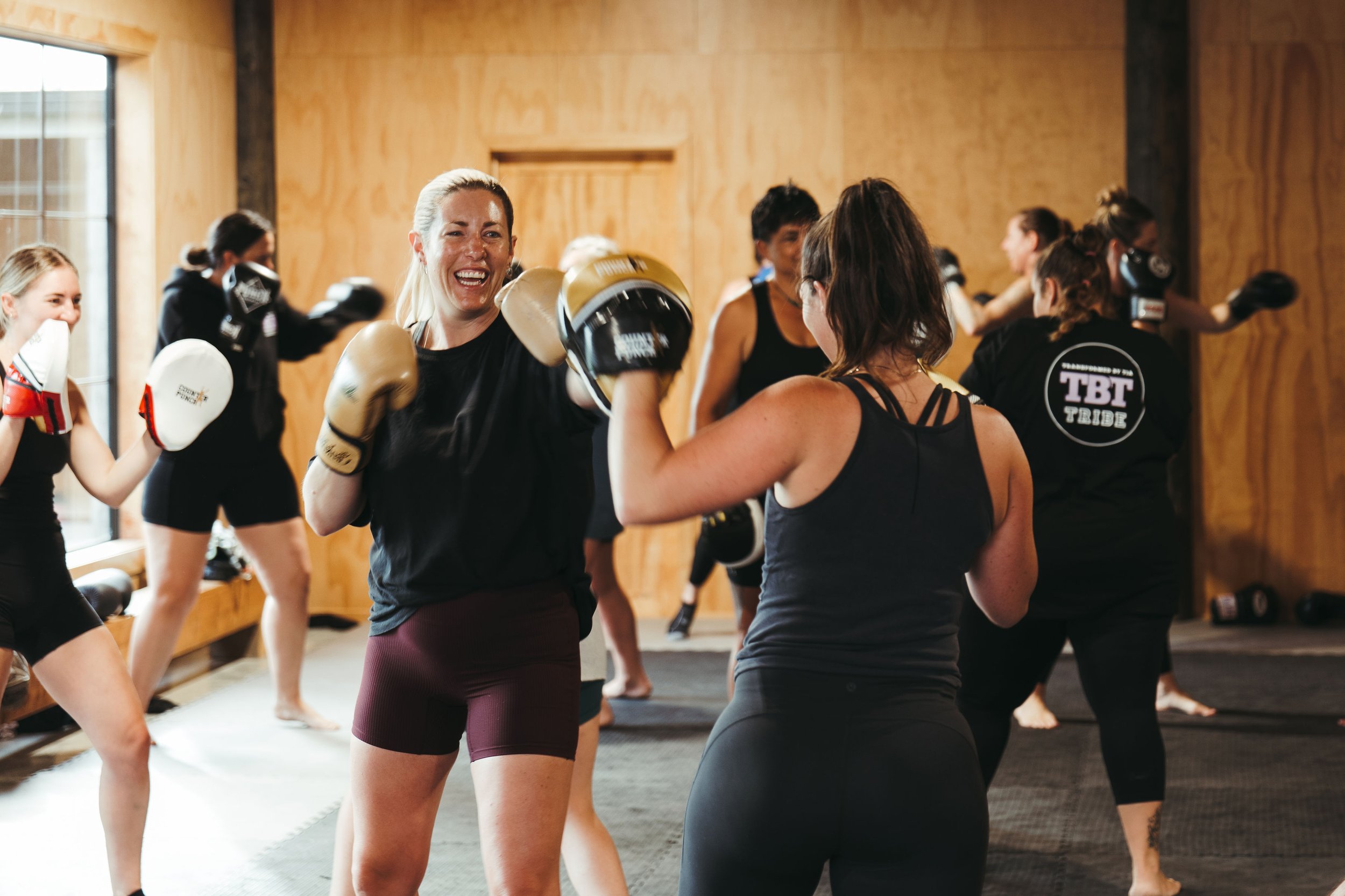 Women participating in a boxing fitness class, wearing boxing gloves and engaging in training exercises in a wooden-floored gym.