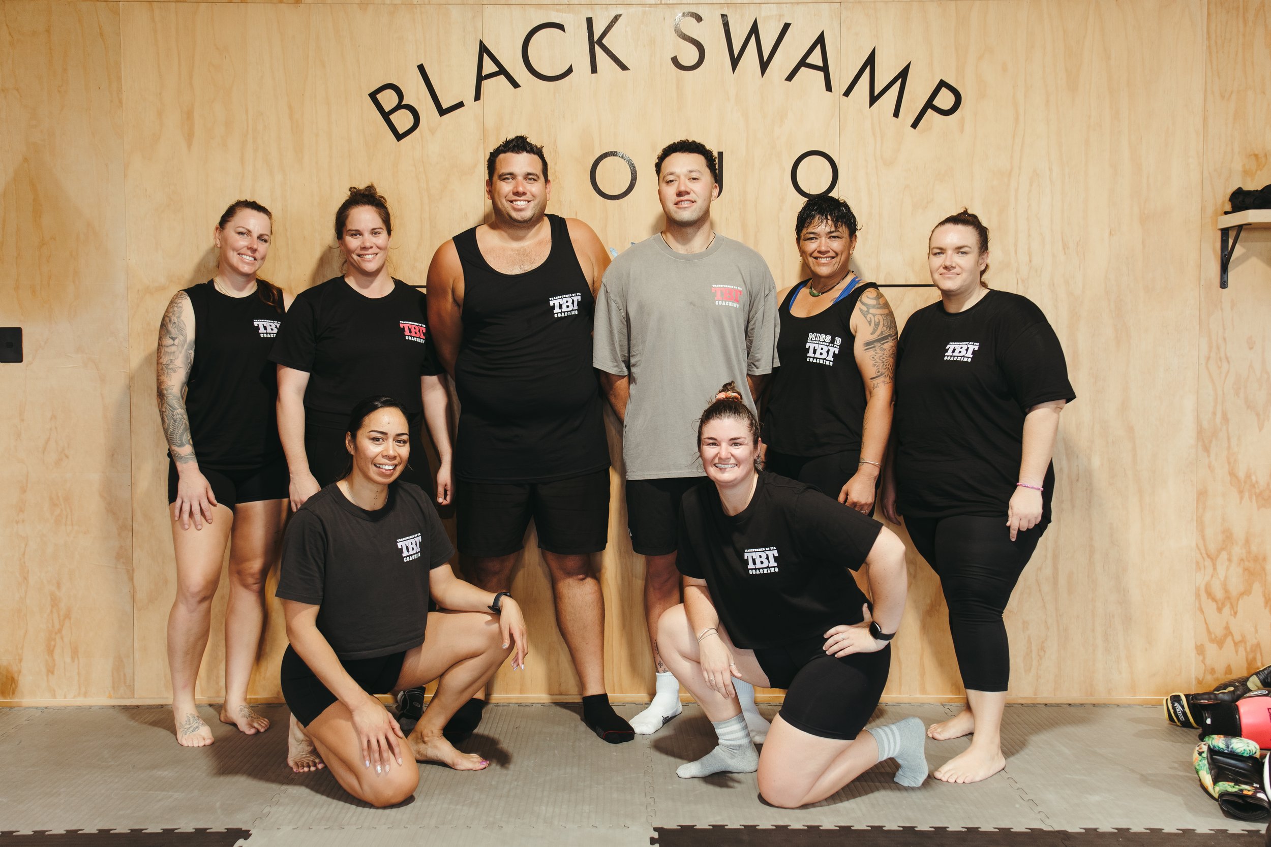 Group of eight diverse people posing in a fitness studio, with some kneeling and others standing, in front of a wooden wall with the words "BLACK SWAMP" written on it. Some are wearing matching workout shirts, and others are in casual athletic wear.
