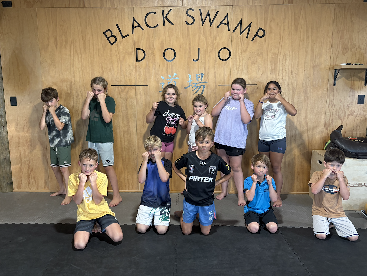 Group of children and a teenager at a martial arts dojo, standing in fighting stances, in front of a wooden wall with the words 'Black Swamp Dojo' displayed.