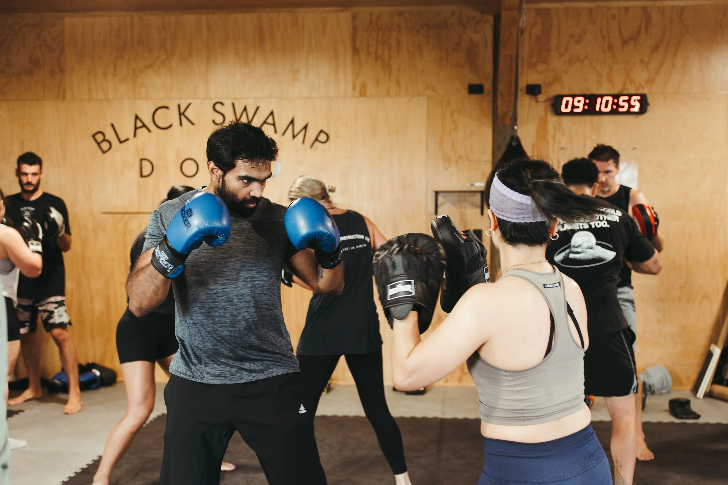 People practicing boxing or martial arts in a gym at Black Swamp dojo, with a wall-mounted digital clock showing 09:10:55.
