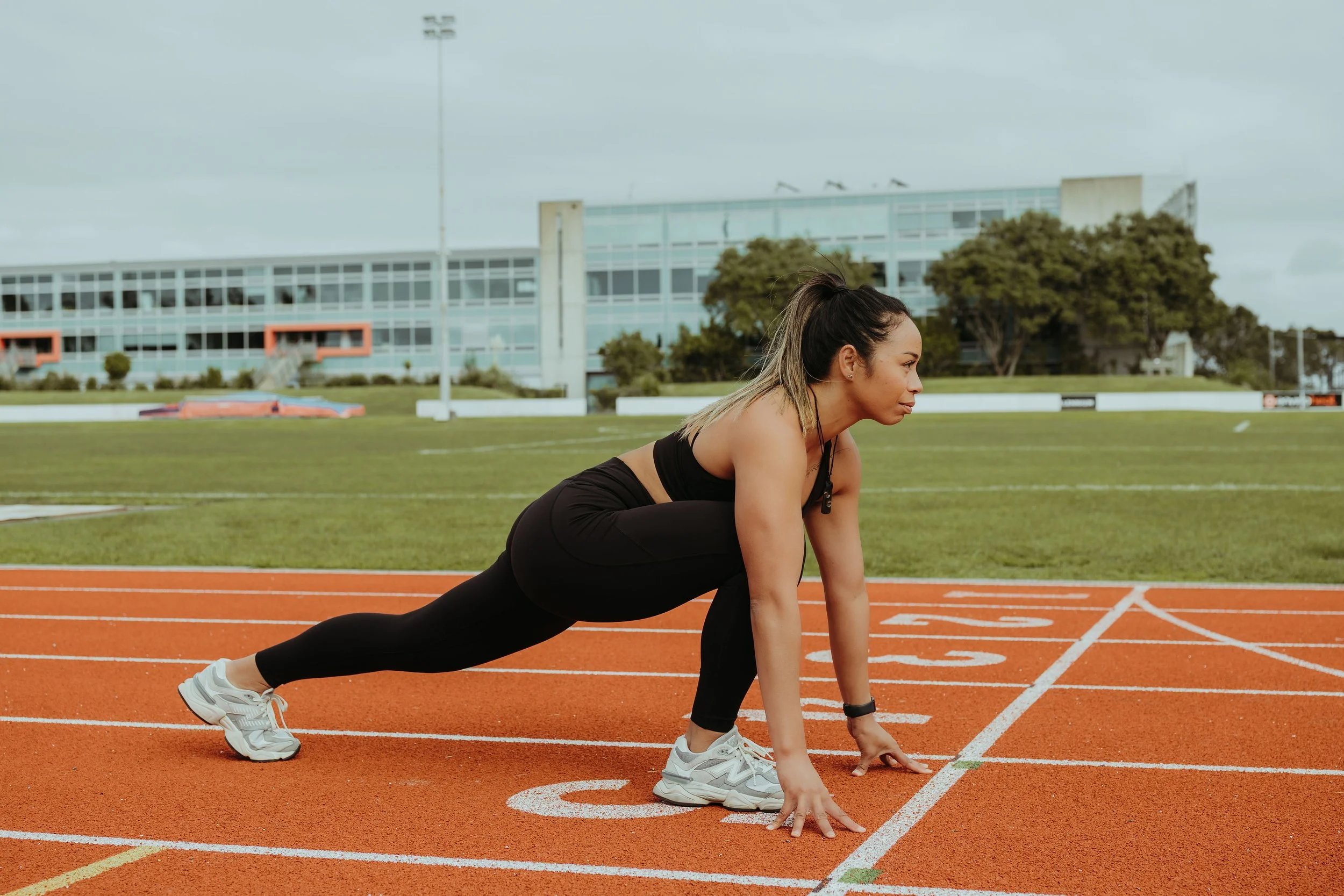 A woman in athletic clothing preparing to run on a track, with a large modern building and trees in the background.