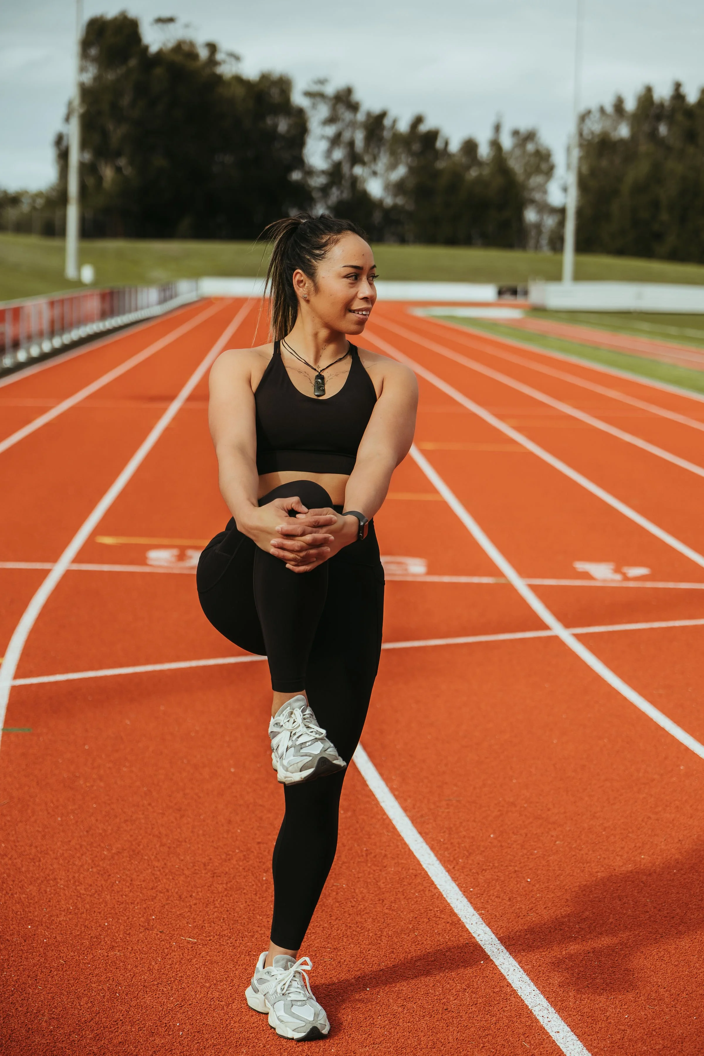 A woman in athletic clothing stretching on a red outdoor running track with trees and light poles in the background.