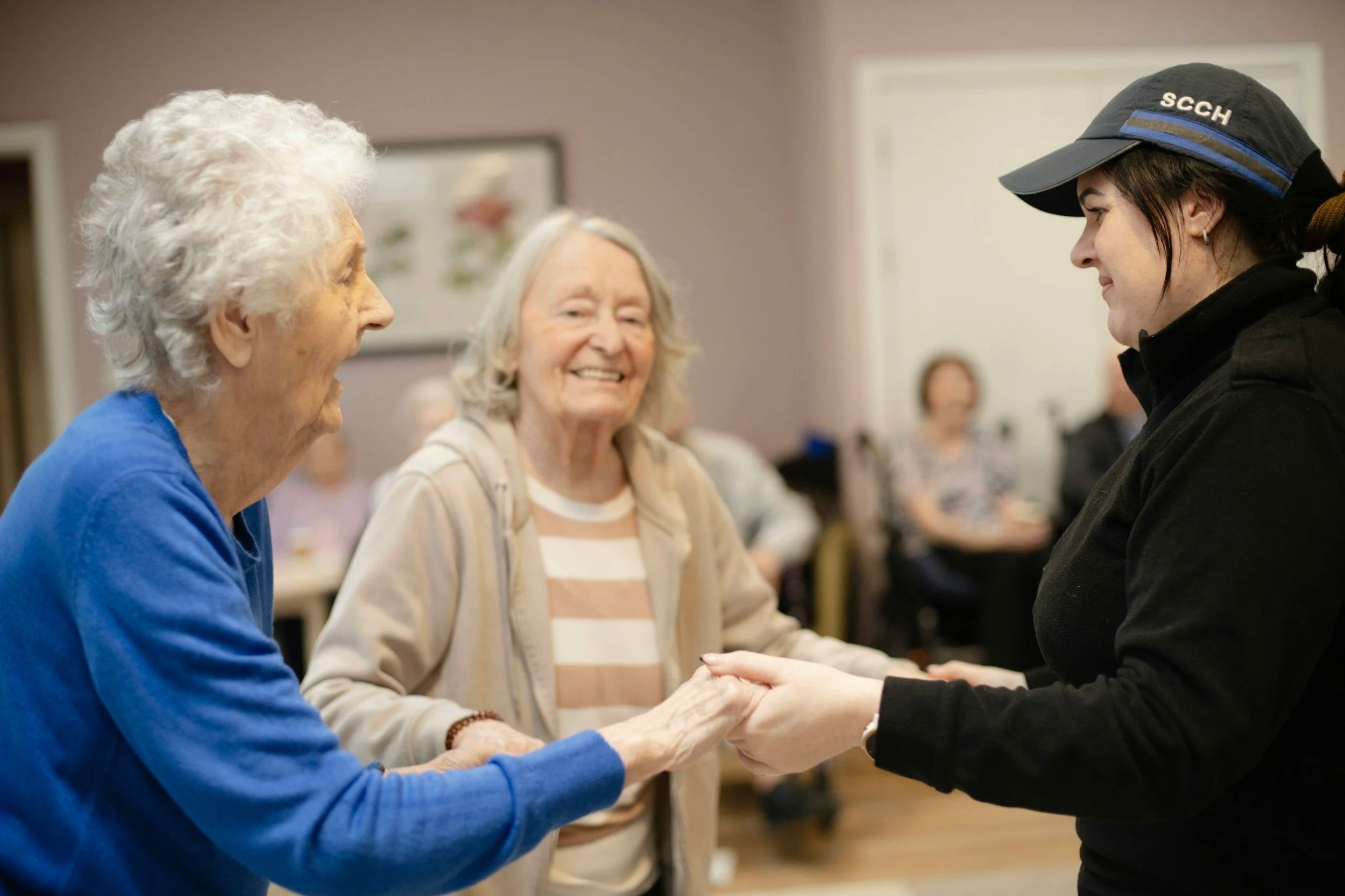 A caregiver in a uniform with 'SCCH' on her hat is holding hands and smiling at two elderly women, who are also smiling and engaging with the caregiver, in a communal room.