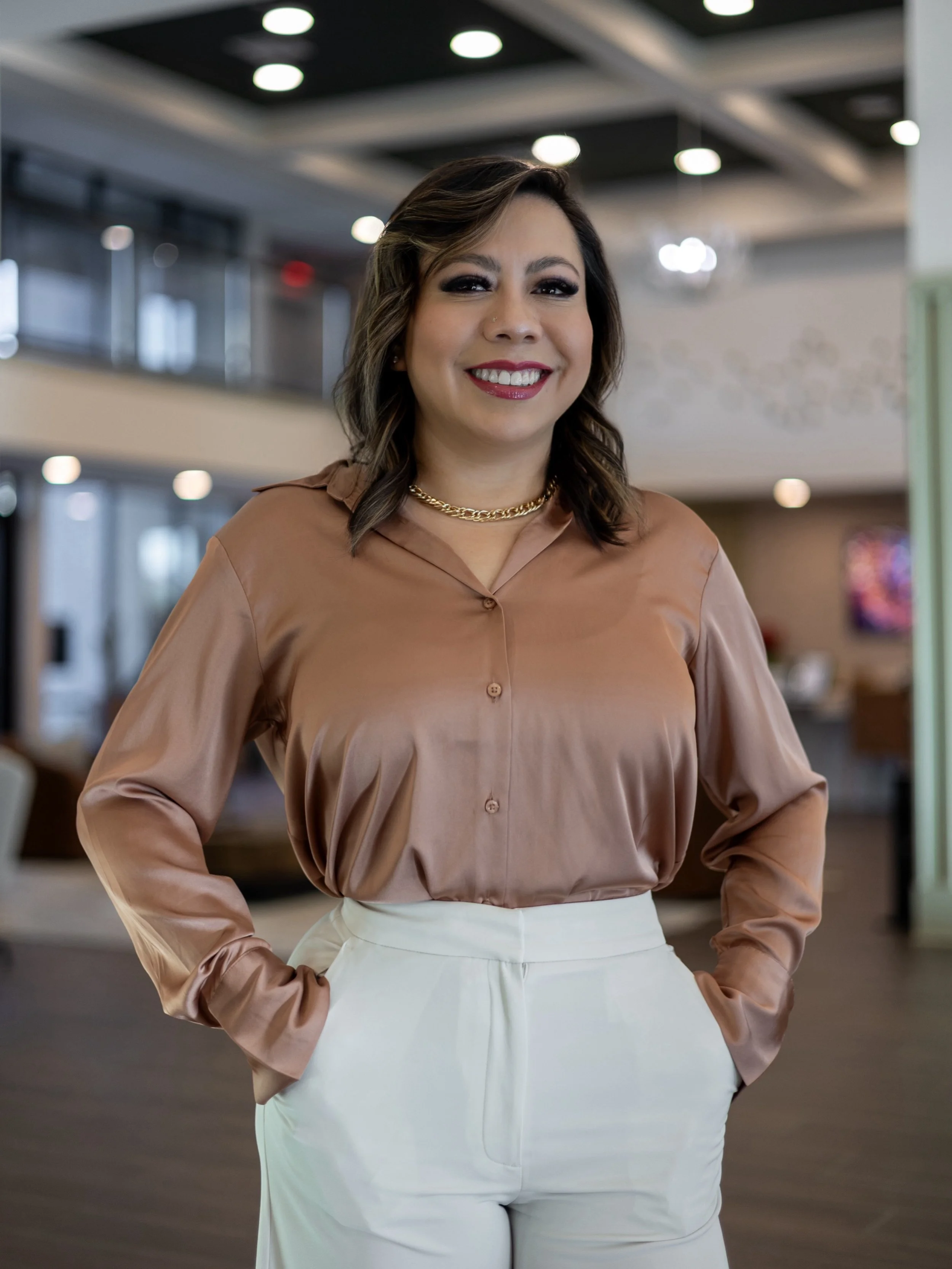 A woman with shoulder-length brown hair, wearing a shiny bronze-colored blouse, white pants, a gold chain necklace, smiling in an indoor space with modern decor.
