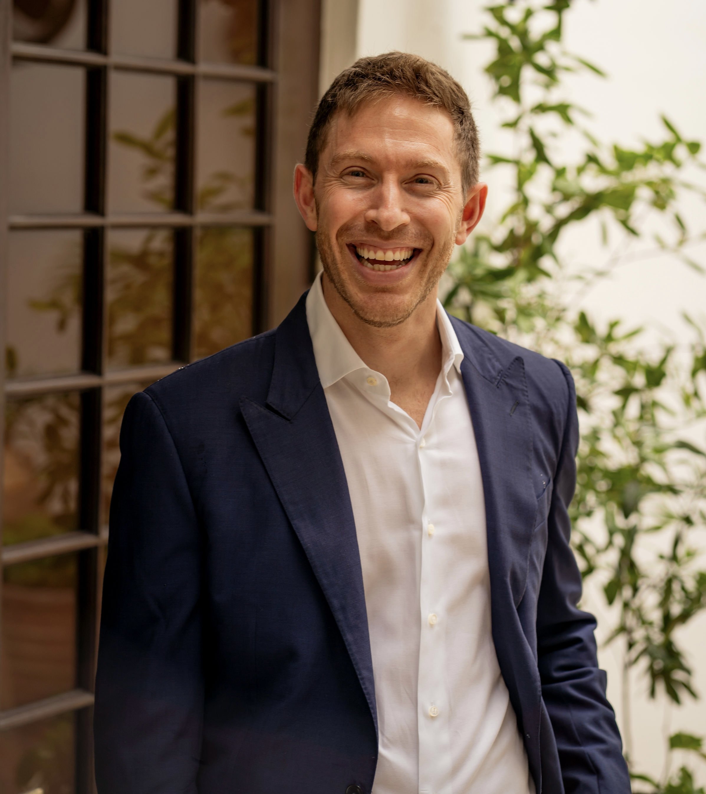 Smiling man in a dark jacket and white shirt, standing outdoors with greenery and a window behind him.