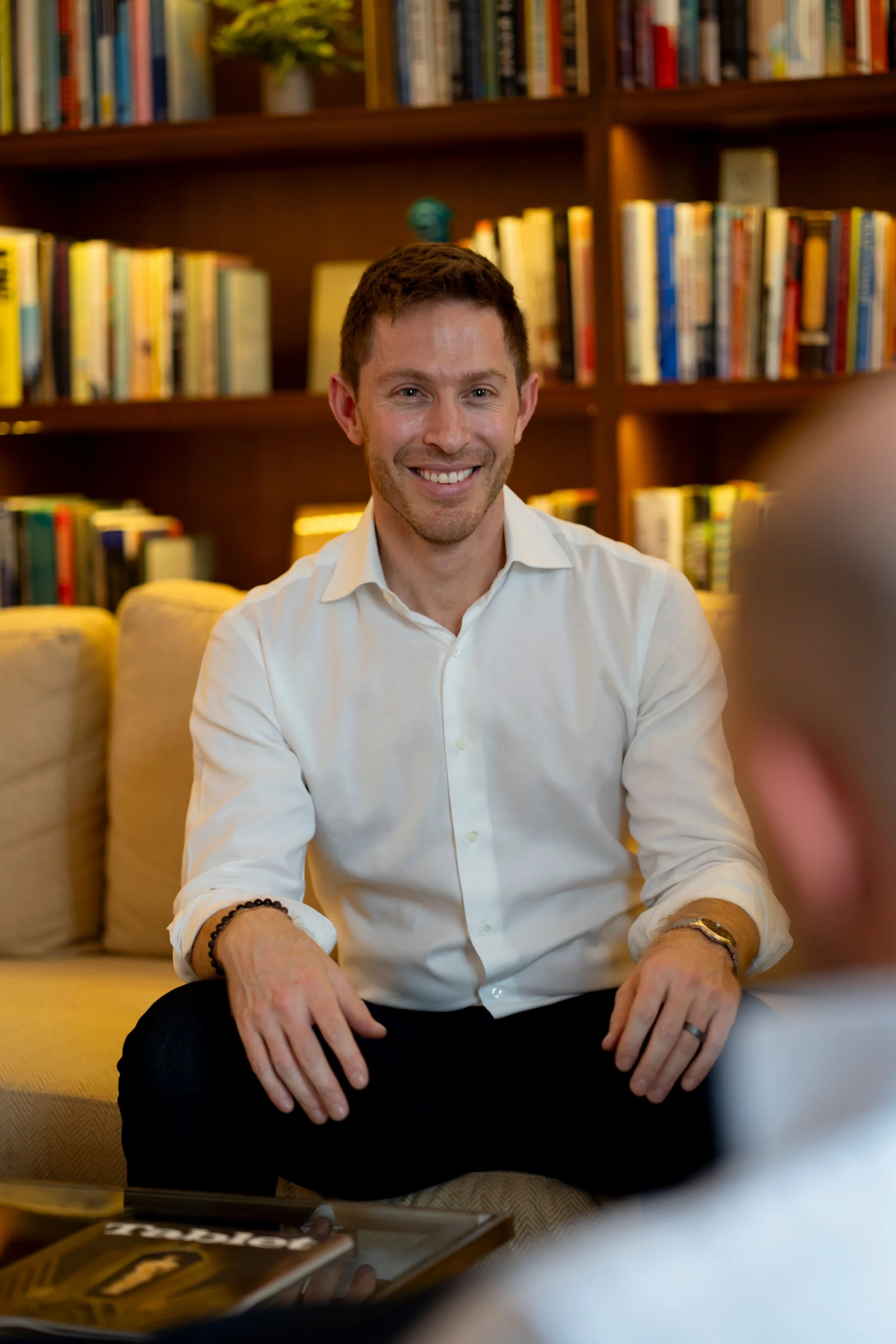 A smiling man with short dark hair, wearing a white button-up shirt, sitting on a beige couch in a room with wooden bookshelves filled with colorful books behind him.