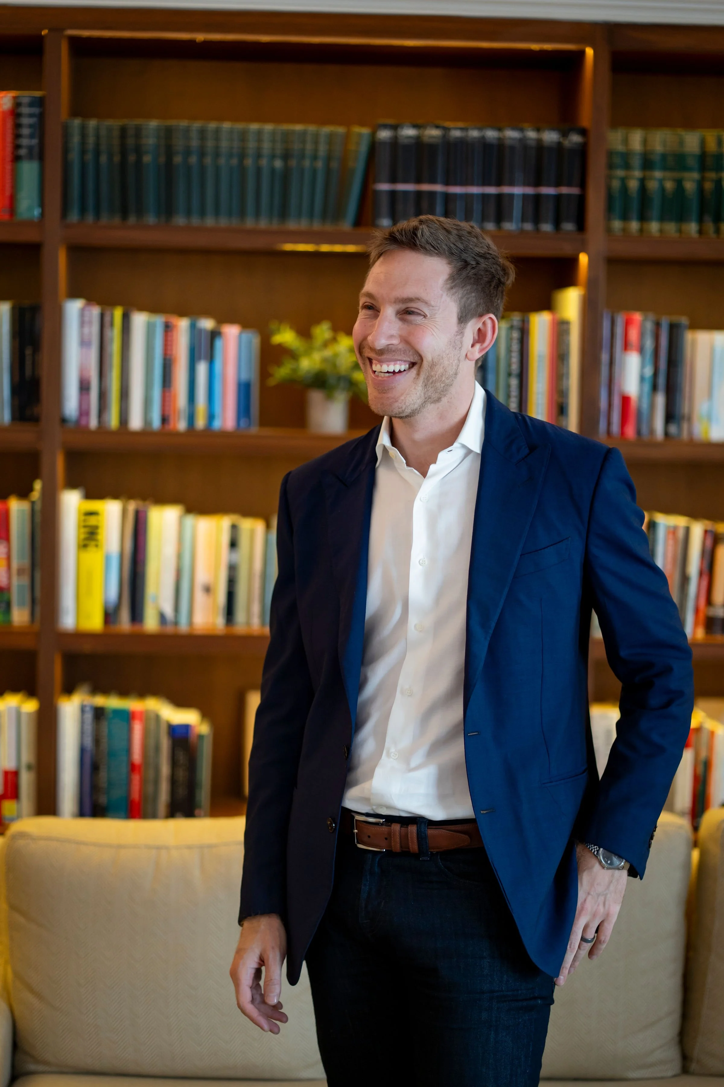 A man in a navy blazer and white shirt smiling, standing in front of a wooden bookshelf filled with books, with a beige sofa in the foreground.