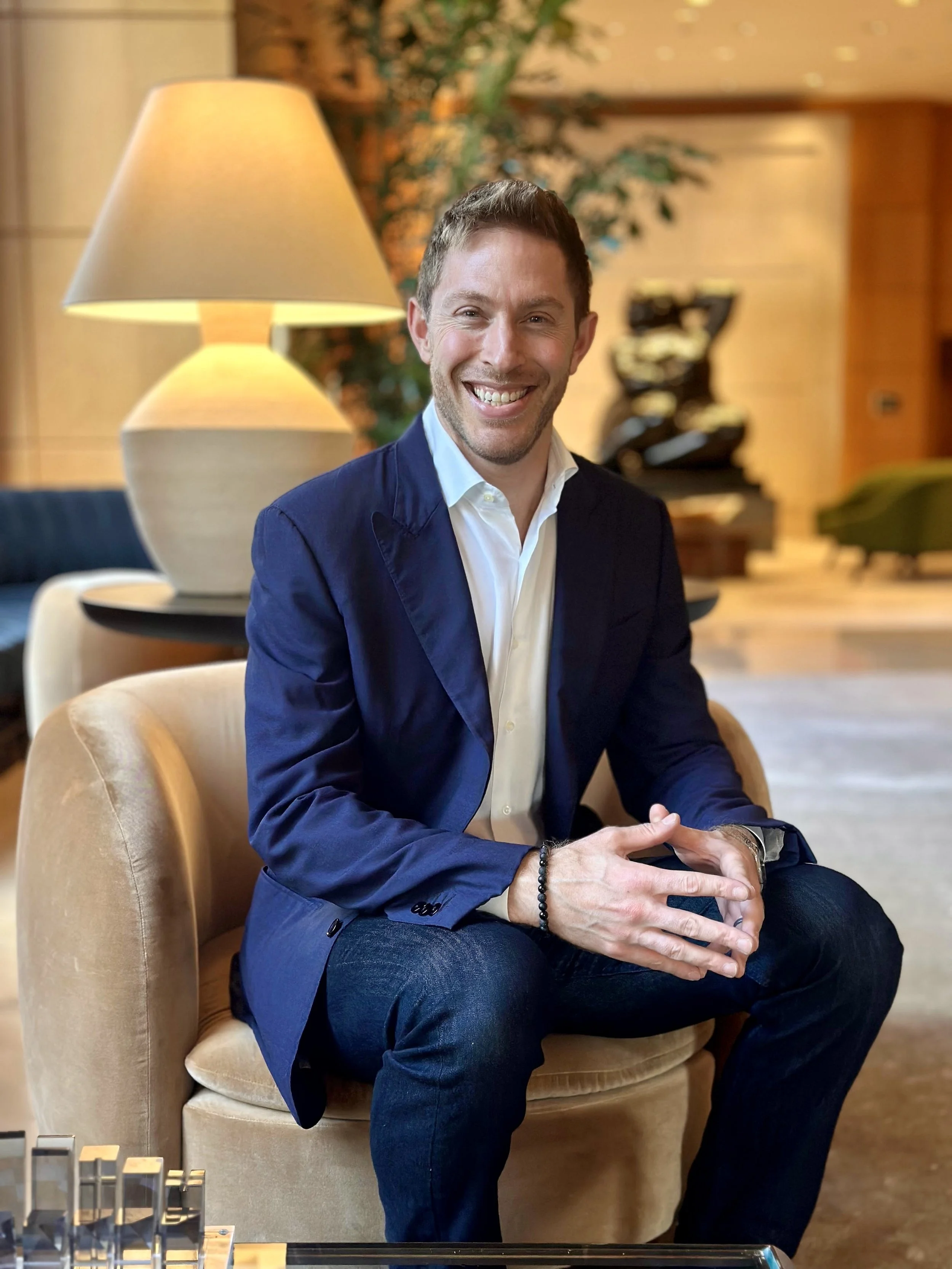 A smiling man in a navy blazer and white shirt sitting on a beige armchair in a warmly lit room with wooden walls, a lamp, and modern art in the background.