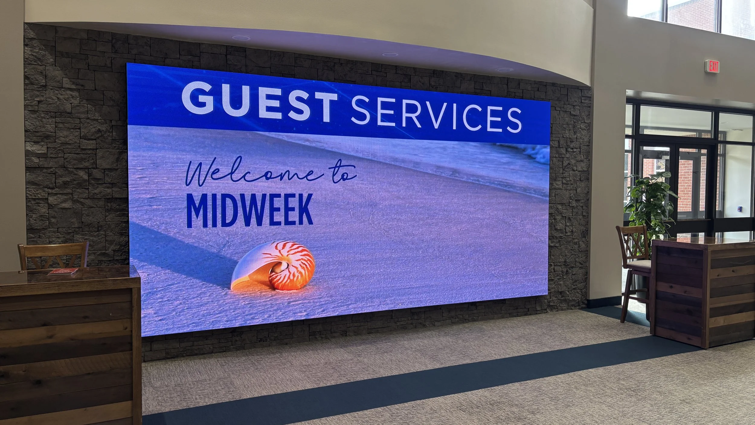 Indoor digital display welcomes guests to Midweek guest services with a beach scene, including sand and a seashell, in a modern lobby area with wooden furnishings and potted plants.