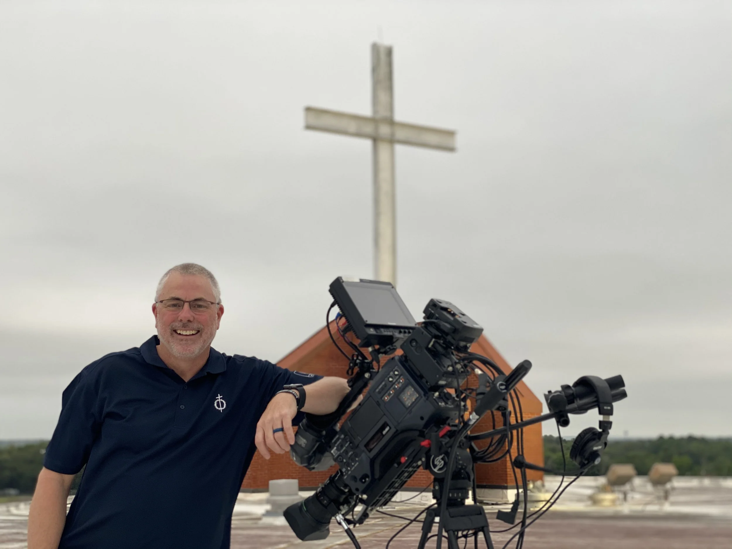 A man with glasses smiling and dressed in a dark polo shirt stands next to a large professional camera on a tripod, with a church steeple and cross behind him in an overcast sky.
