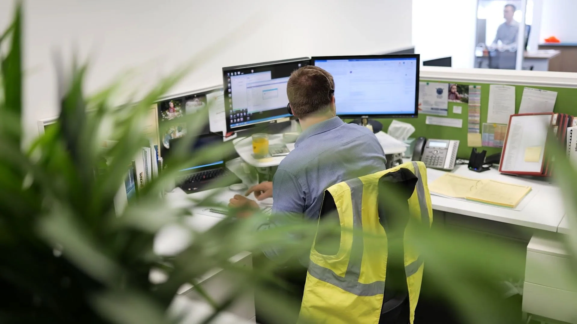 Behind-the-shoulder view of an office worker using dual monitors at a desk, with a yellow high-visibility safety vest hanging on his chair.