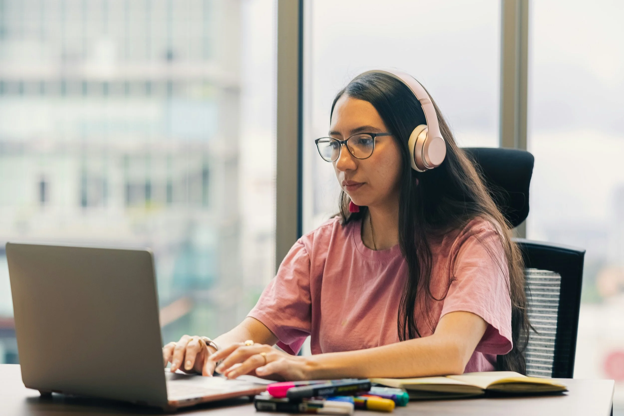 A focused woman wearing glasses and headphones typing on her laptop at a desk in front of a high-rise office window.