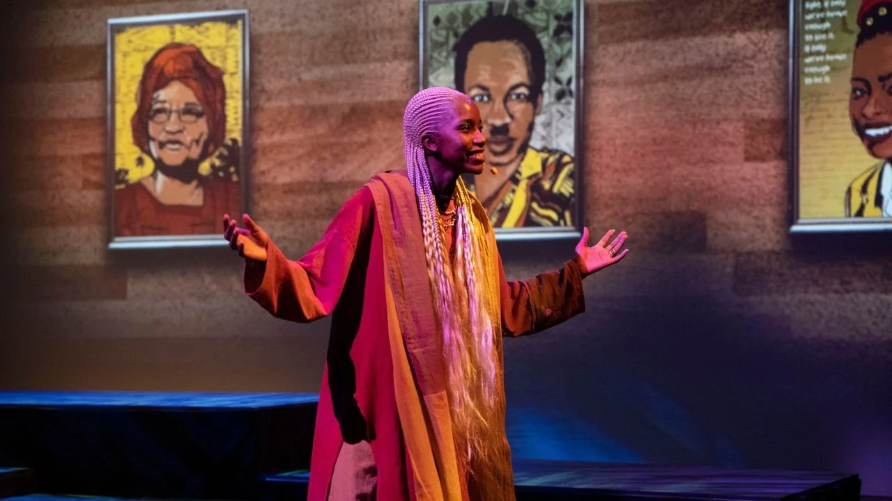 Performer Vivana Luzochimana onstage during a production of The Deep North, gesturing with open arms in front of illustrated backdrop portraits.