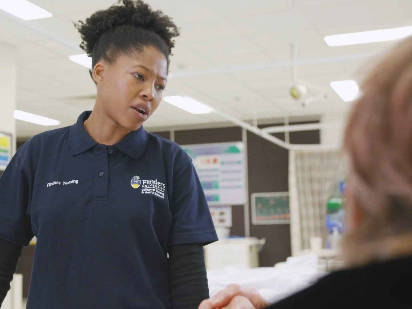 Still from an educational video production showing a healthcare worker mid-conversation during a simulated nursing training scenario.