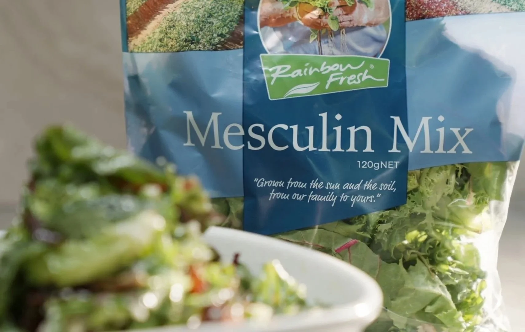 Close-up of a Rainbow Fresh Mesculin Mix salad bag with a blurred bowl of fresh salad greens in the foreground.