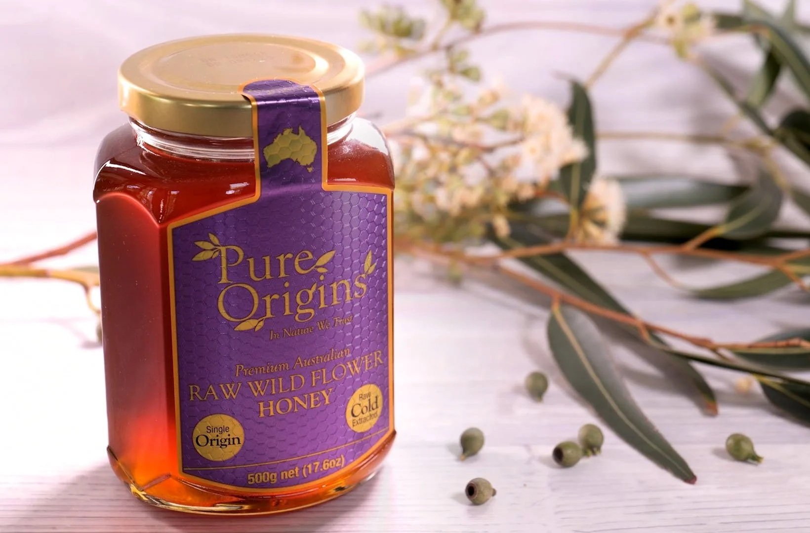 Pure Origins Australian raw wildflower honey 500g glass jar. Premium single origin raw cold-extracted honey from Australia, styled on a light wooden background with native eucalyptus leaves and blossoms.
