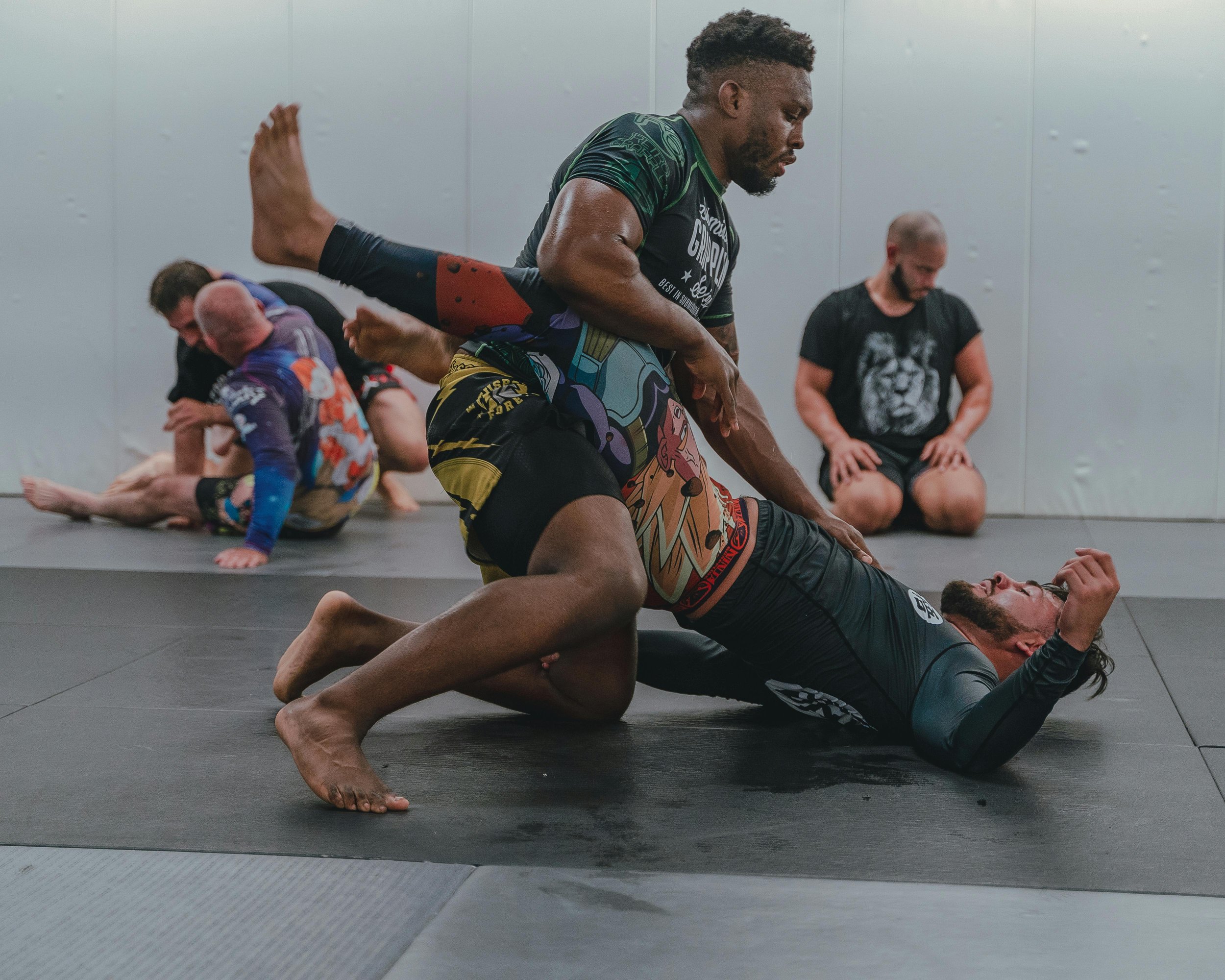 Two men practicing Brazilian Jiu-Jitsu on a black mat in a gym, with one man on top of the other, both in grappling positions.