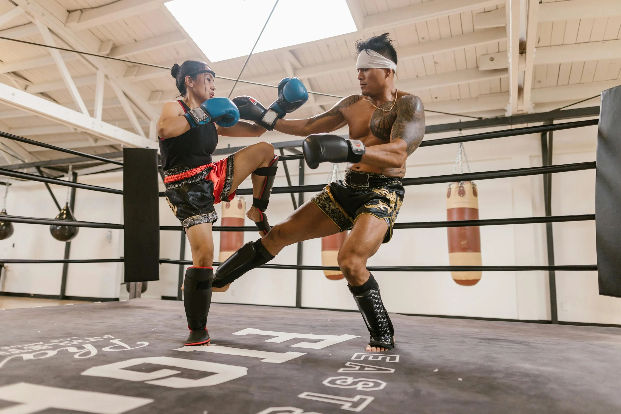 Two fighters sparring in a boxing ring, one delivering a punch and the other blocking, both wearing boxing gloves and shorts, with punching bags hanging in the background.