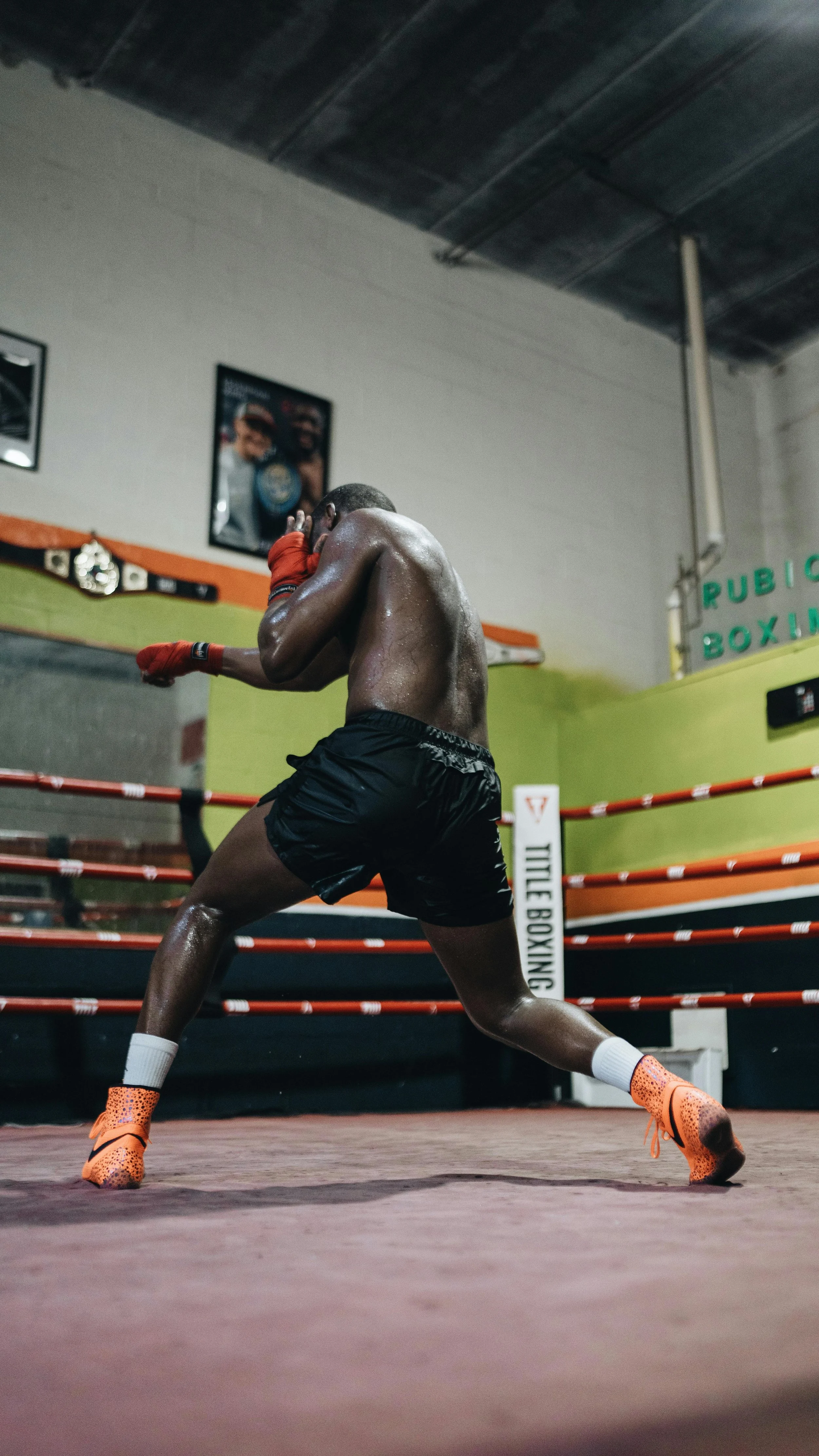 A male boxer training in a boxing gym, wearing black shorts, orange boxing shoes, and red hand wraps, throwing a punch in a boxing ring.