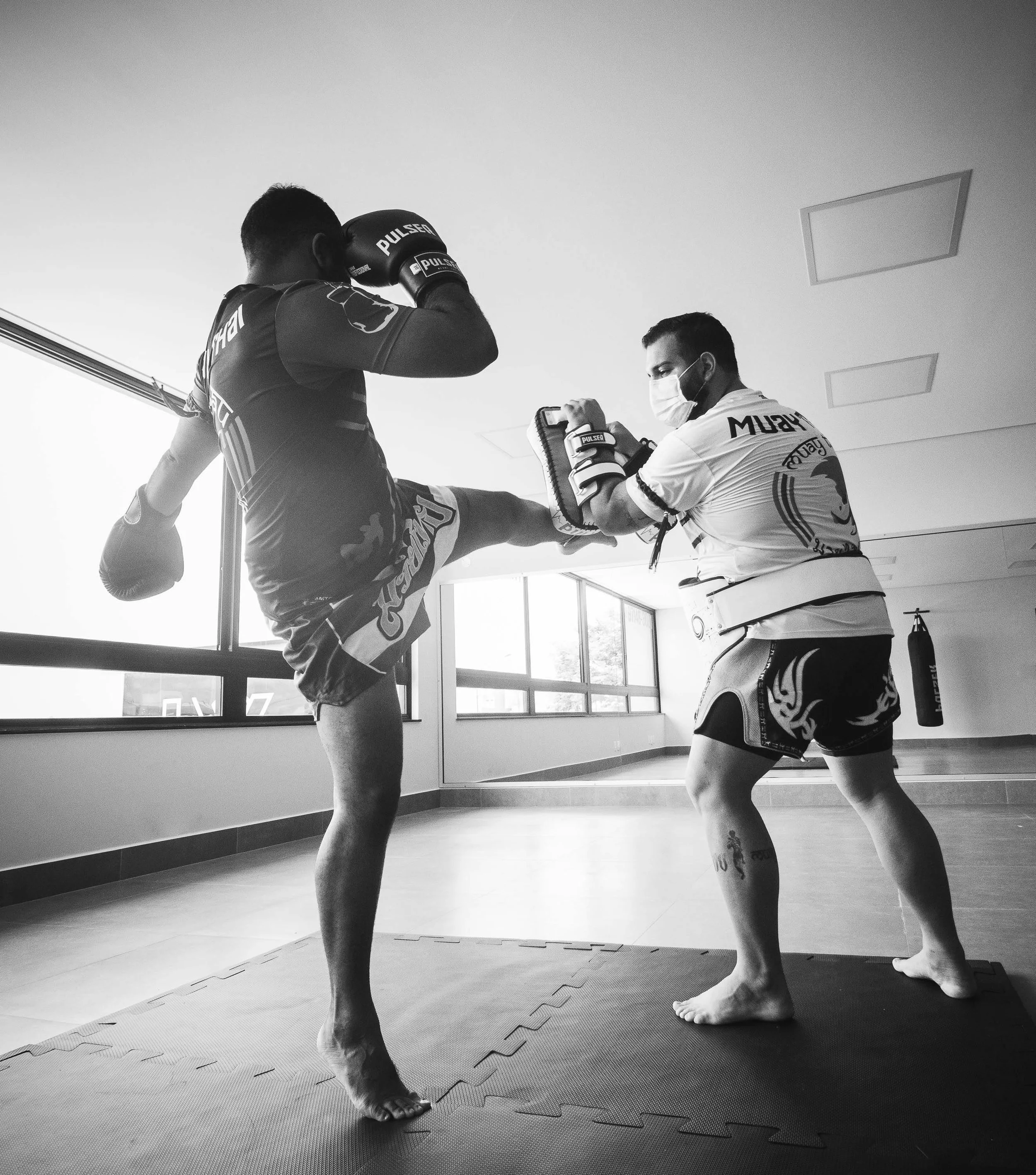 Two martial arts practitioners practicing kickboxing or mixed martial arts in a gym. One person is wearing boxing gloves and a team uniform, and is executing a kick towards the other who is wearing a protective school and raising their arms in a defensive stance. Both are standing on a padded mat in front of large windows, with a punching bag hanging in the background.