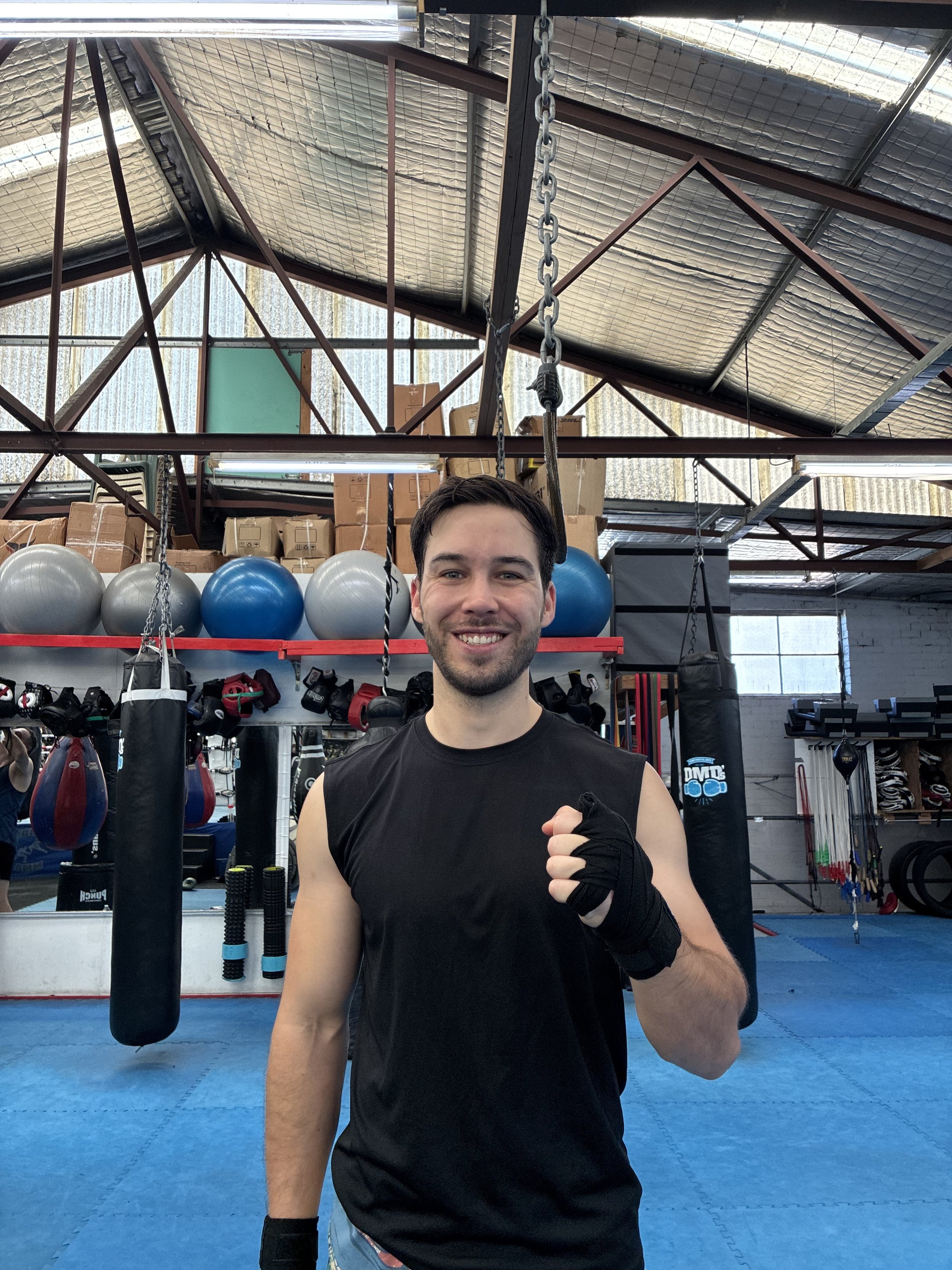 A young man posing with a fist raised in a boxing gym. He is wearing a black athletic shirt and boxing gloves, with exercise equipment and boxing bags in the background.