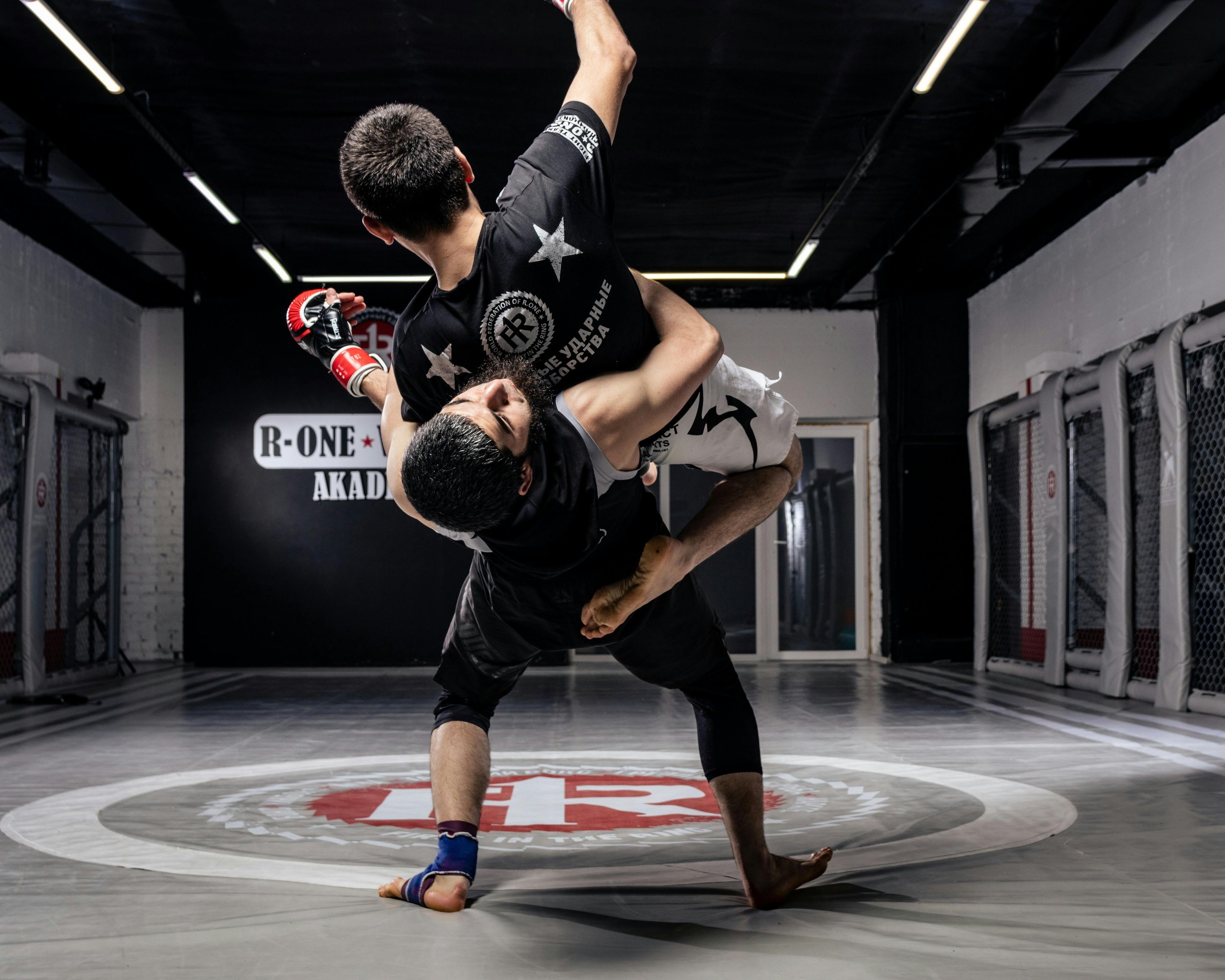 Two men practicing Brazilian jiu-jitsu in a martial arts gym. One man is executing a move on the other, who is on the mat. The gym has a black ceiling, gray walls, and a branding sign that says "R-One Academy" in the background.