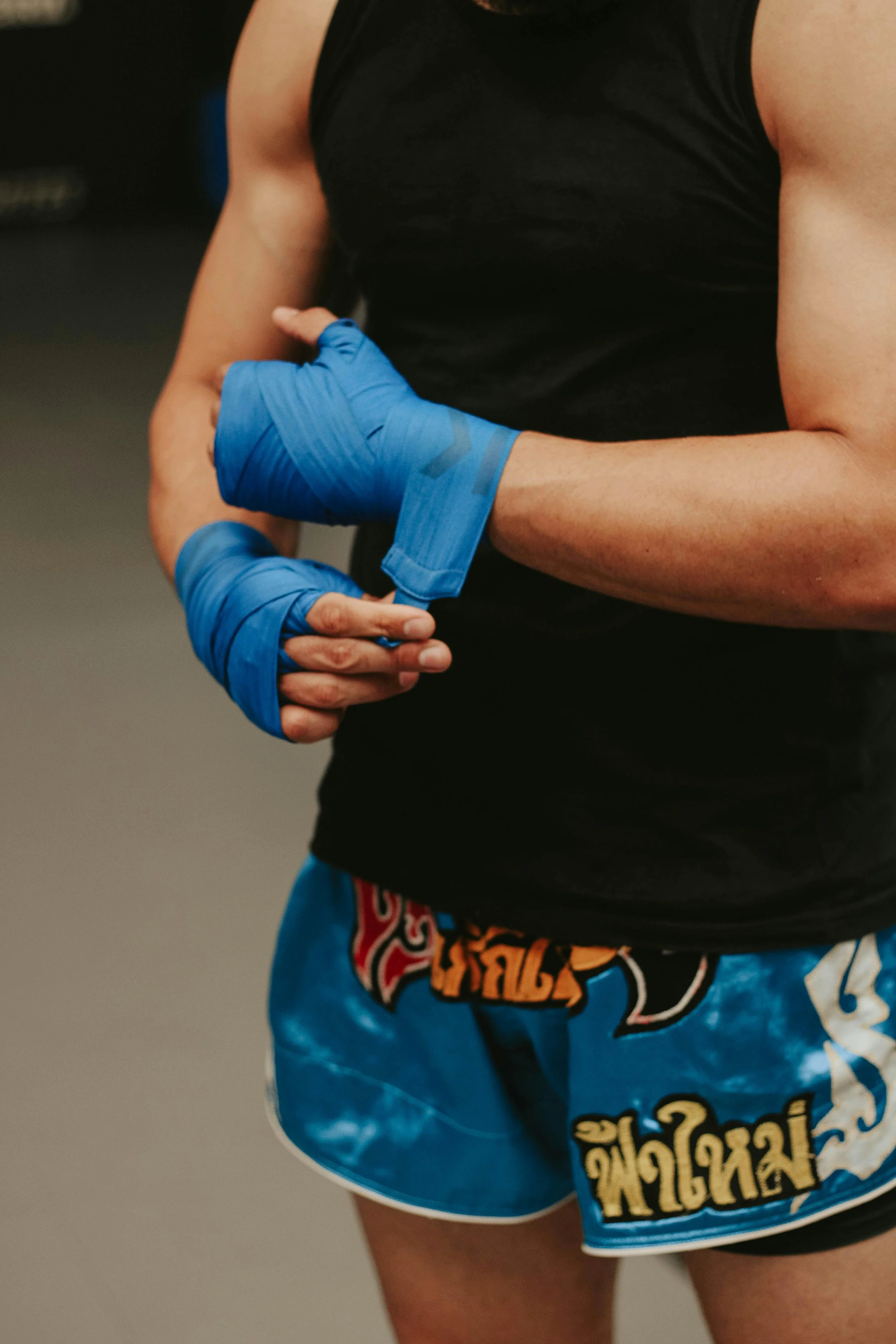 A person wearing black sleeveless shirt and blue shorts with yellow and orange text, wrapping blue hand wraps around their fists, likely preparing for a boxing or martial arts workout.