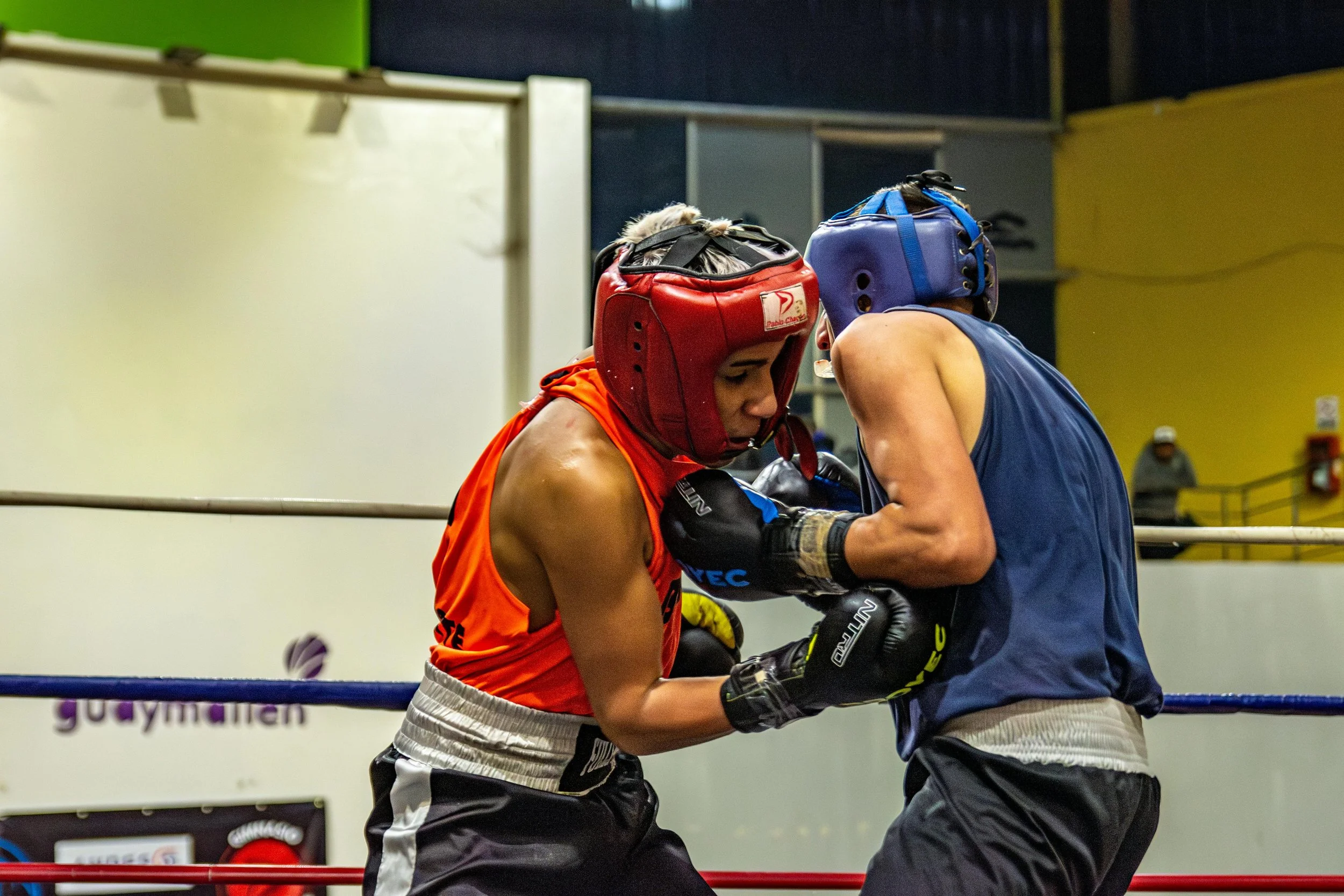 Two male boxers in protective headgear and gloves inside a boxing ring, engaged in a sparring session with their foreheads touching.