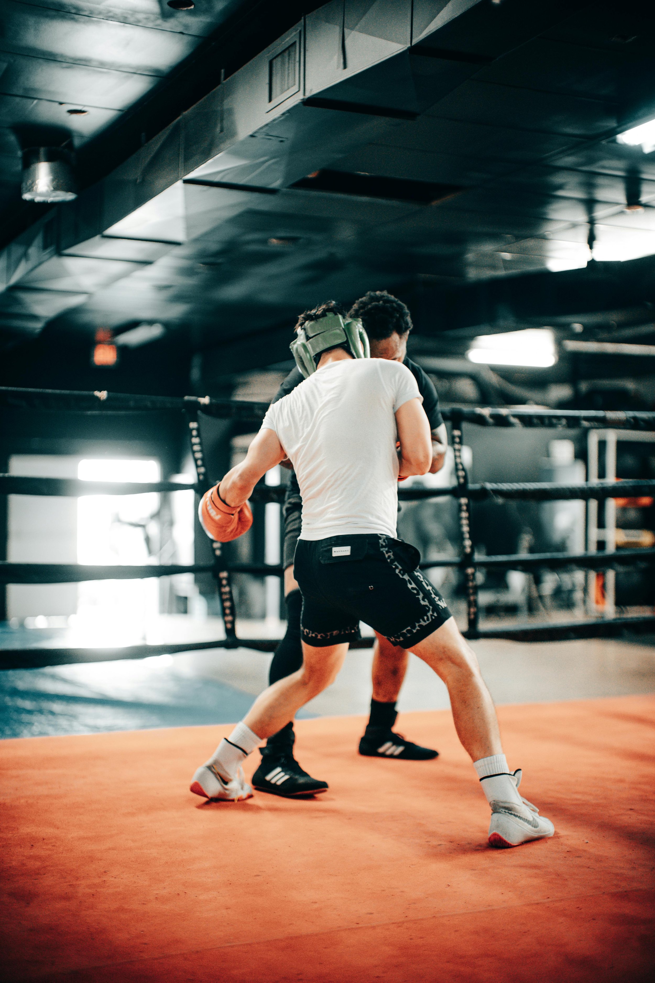 Two men training in a boxing gym, one wearing boxing gloves, practicing fighting moves in a ring with a black padded floor.