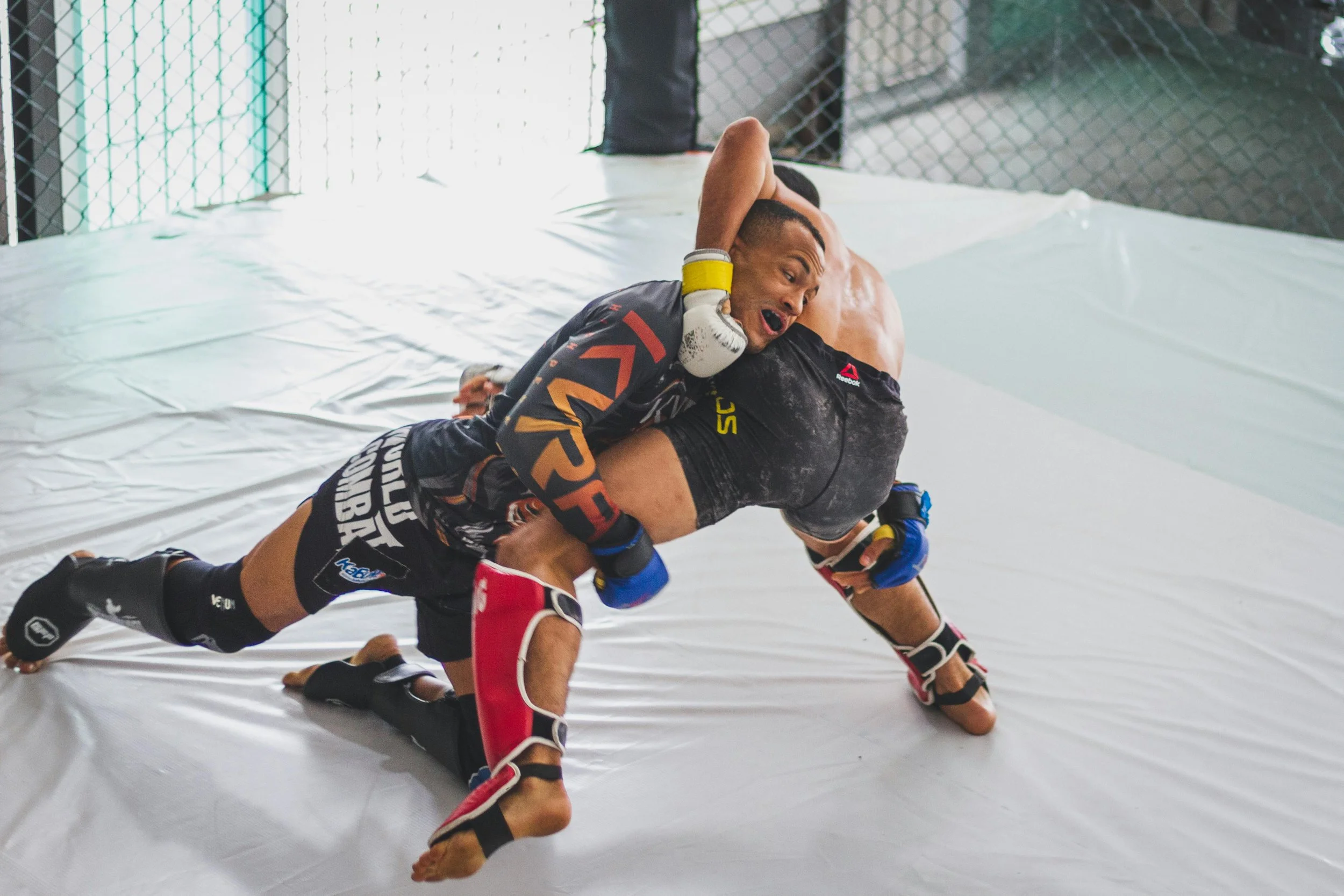 Two mixed martial arts fighters engaged on a mat inside a cage. One fighter is on top attempting a submission move while the other is on the ground trying to escape. Both are wearing shorts, gloves, and protective gear.
