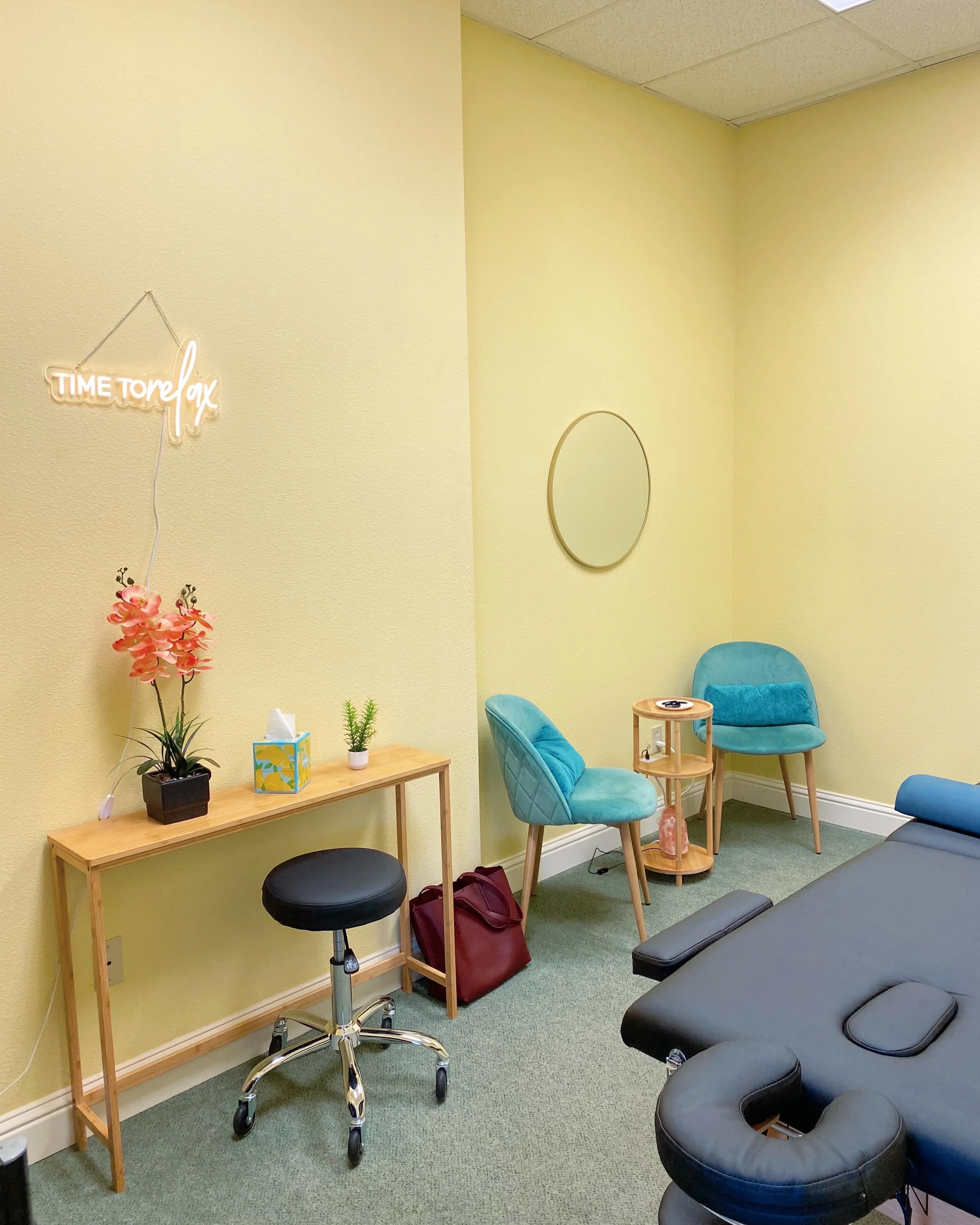 Dr. Dhami's Santa Clara office, with a white neon sign stating "time to relax", a console table with tissues and flowers on top, Dr. Dhami's trusty family medicine spinning stool, 2 teal chairs around a small side table for convos, an exam table