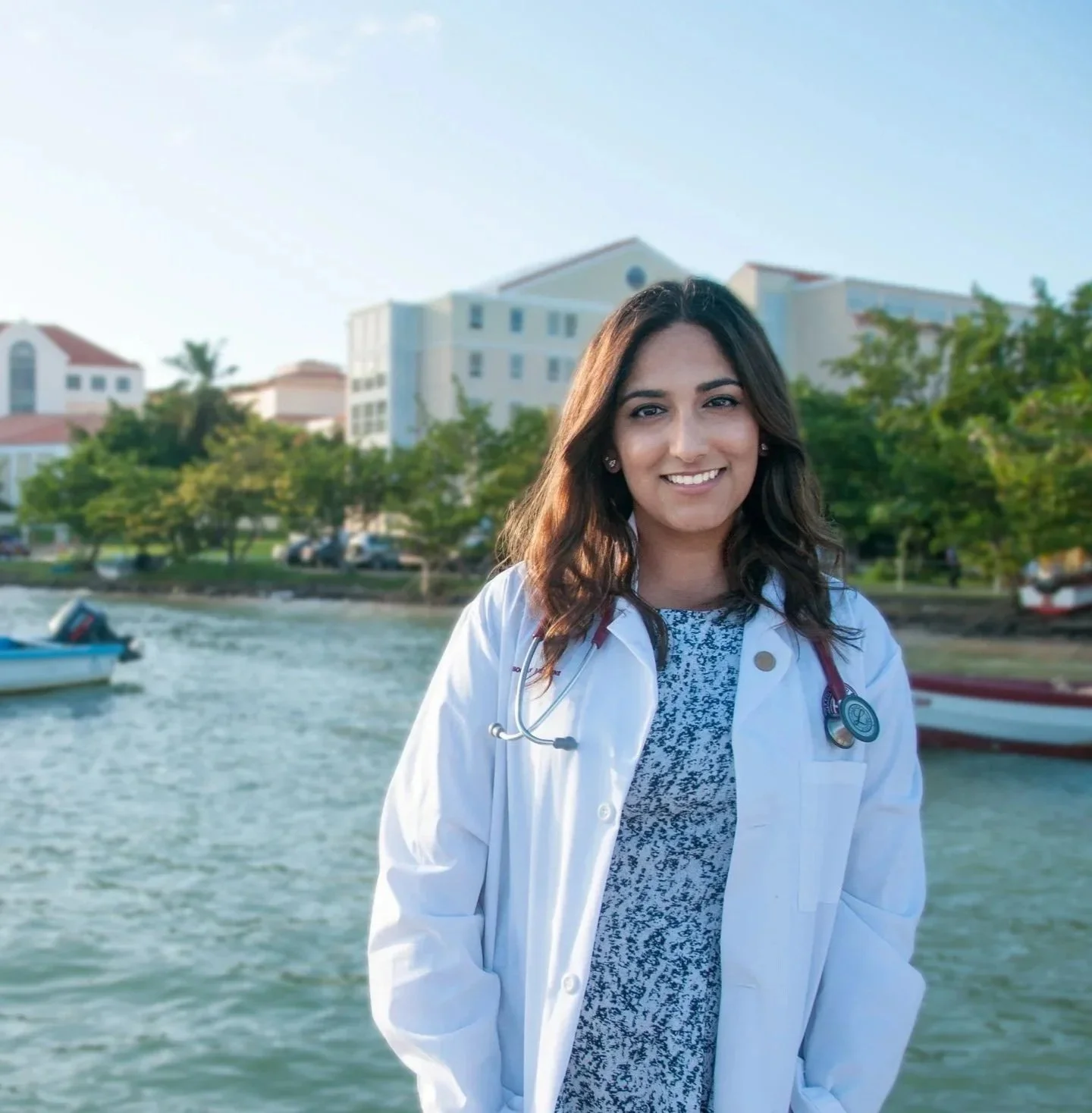 Dr. Rubeena Dhami, MD wearing a physician's white coat and stethoscope, standing in front of St. George's University campus in Grenada