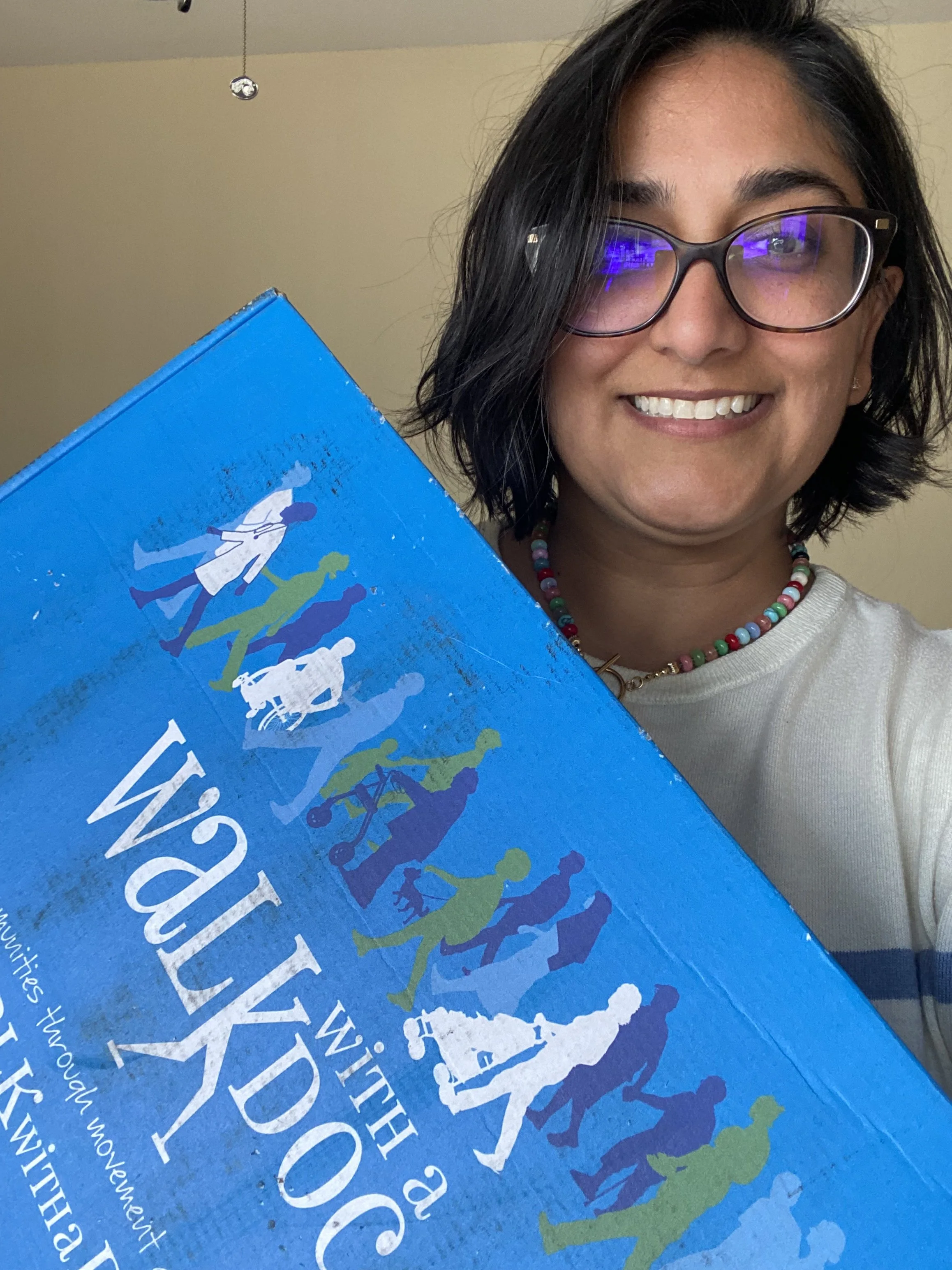 Dr. Dhami with black hair, glasses, and a colorful beaded necklace holding a blue box with illustrated figures and text on the cover reading Walk with a Doc, smiling at the camera. The background is a plain beige wall.