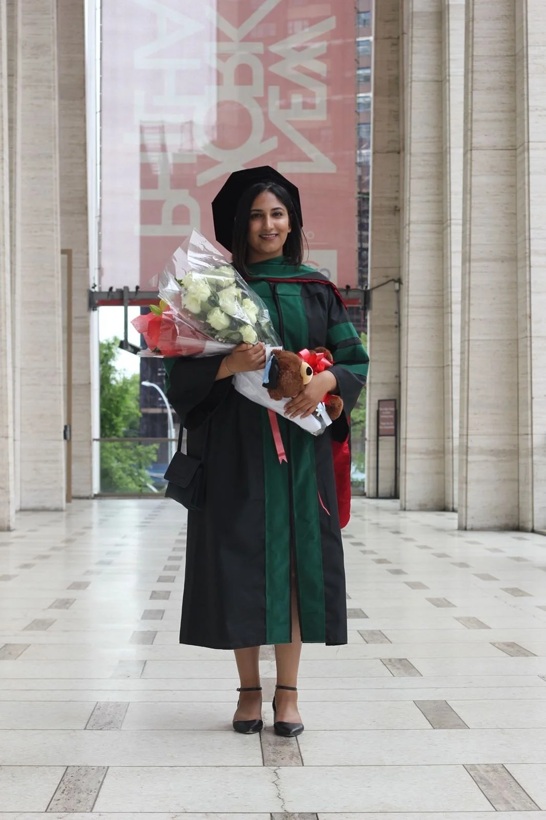 Dr. Rubeena Dhami, MD in her SGU SOM graduation regalia holding a bouquet of flowers and a teddy bear, standing inside Lincoln Center with tall columns and a large banner in the background.