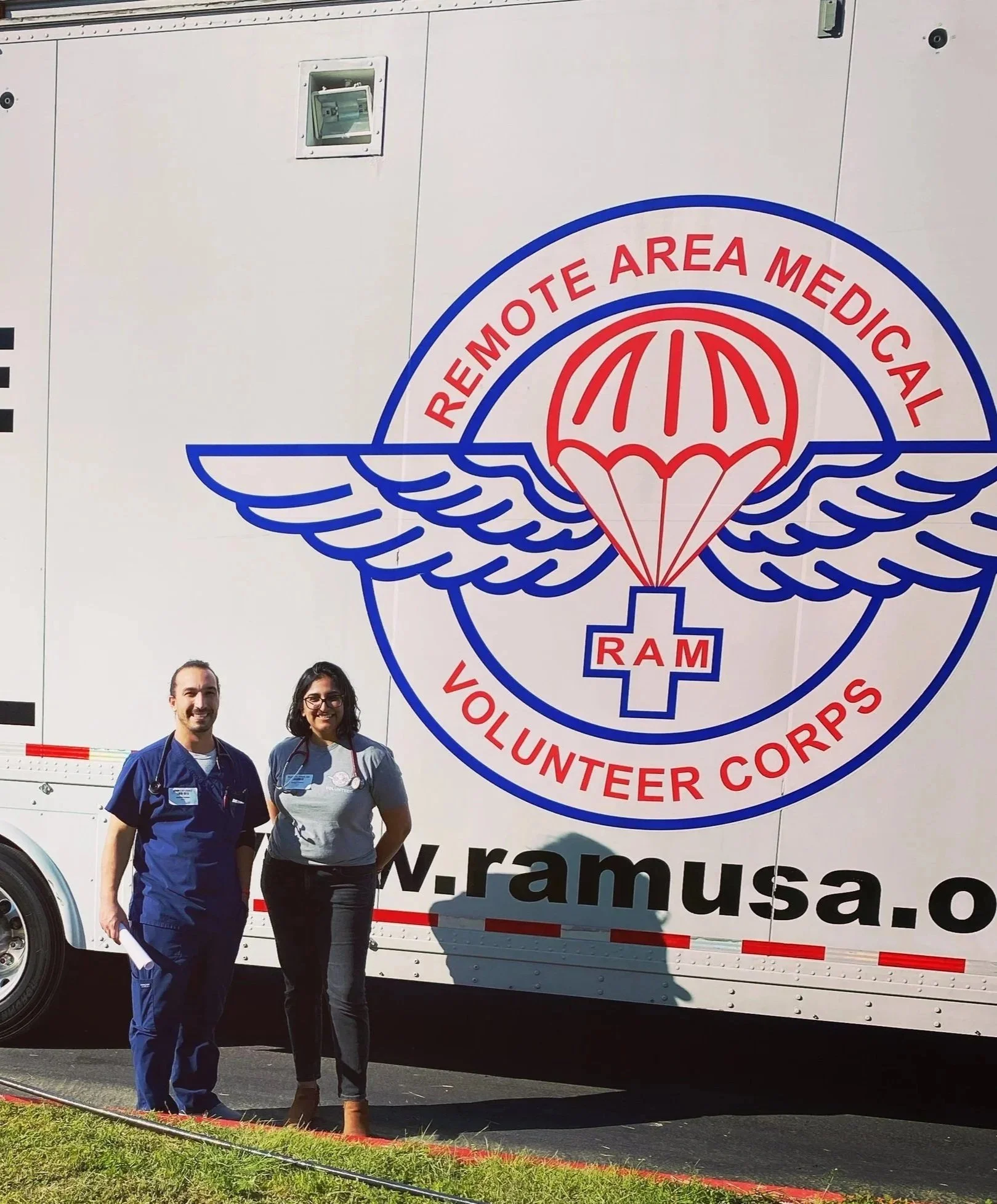 Dr. Dhami and a colleague in front of a mobile Remote Area Medical vehicle with a large logo, smiling at the camera.
